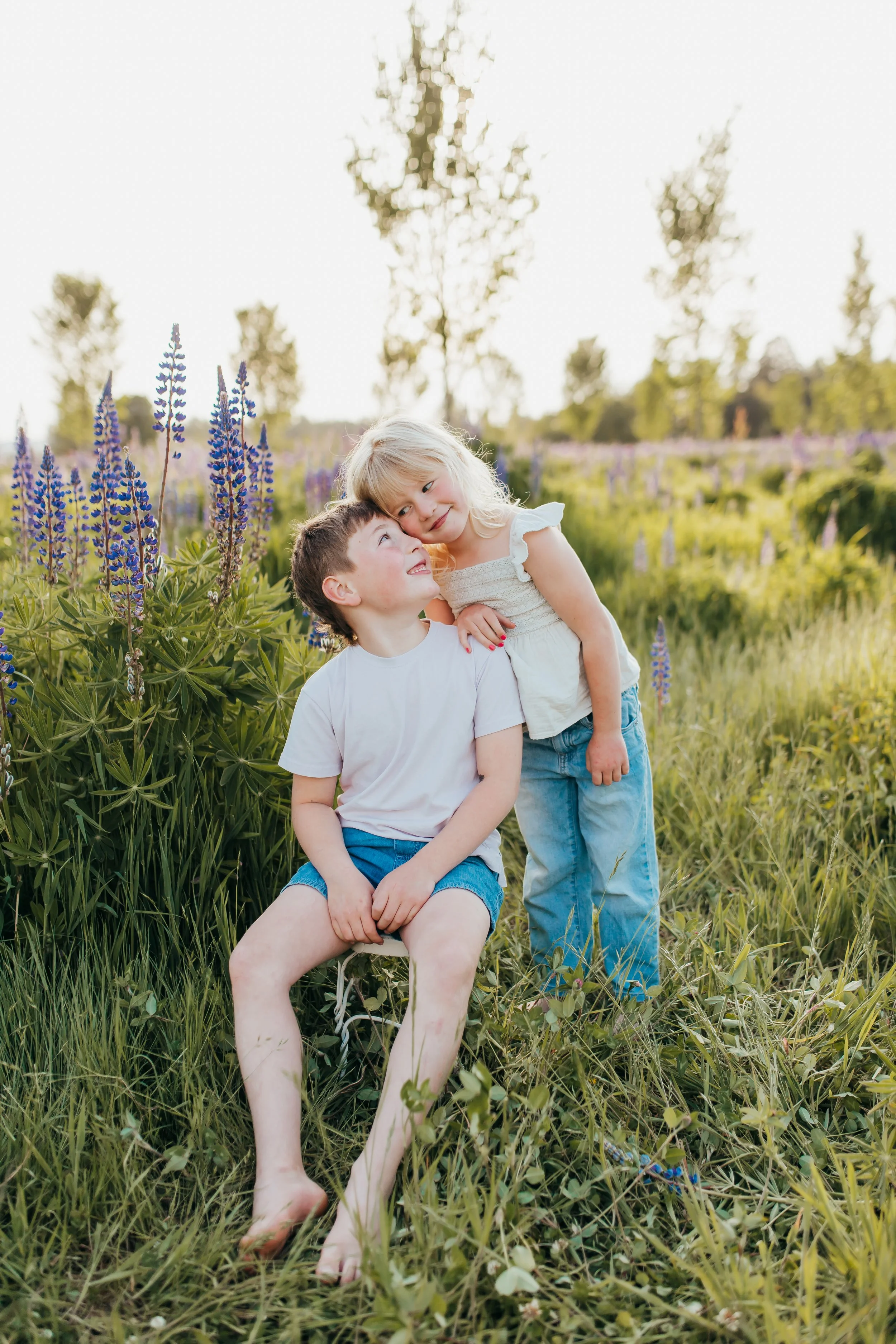 Two children, a boy and a girl, in a field of lupine flowers, sitting and leaning on each other, with trees and a bright sky in the background.