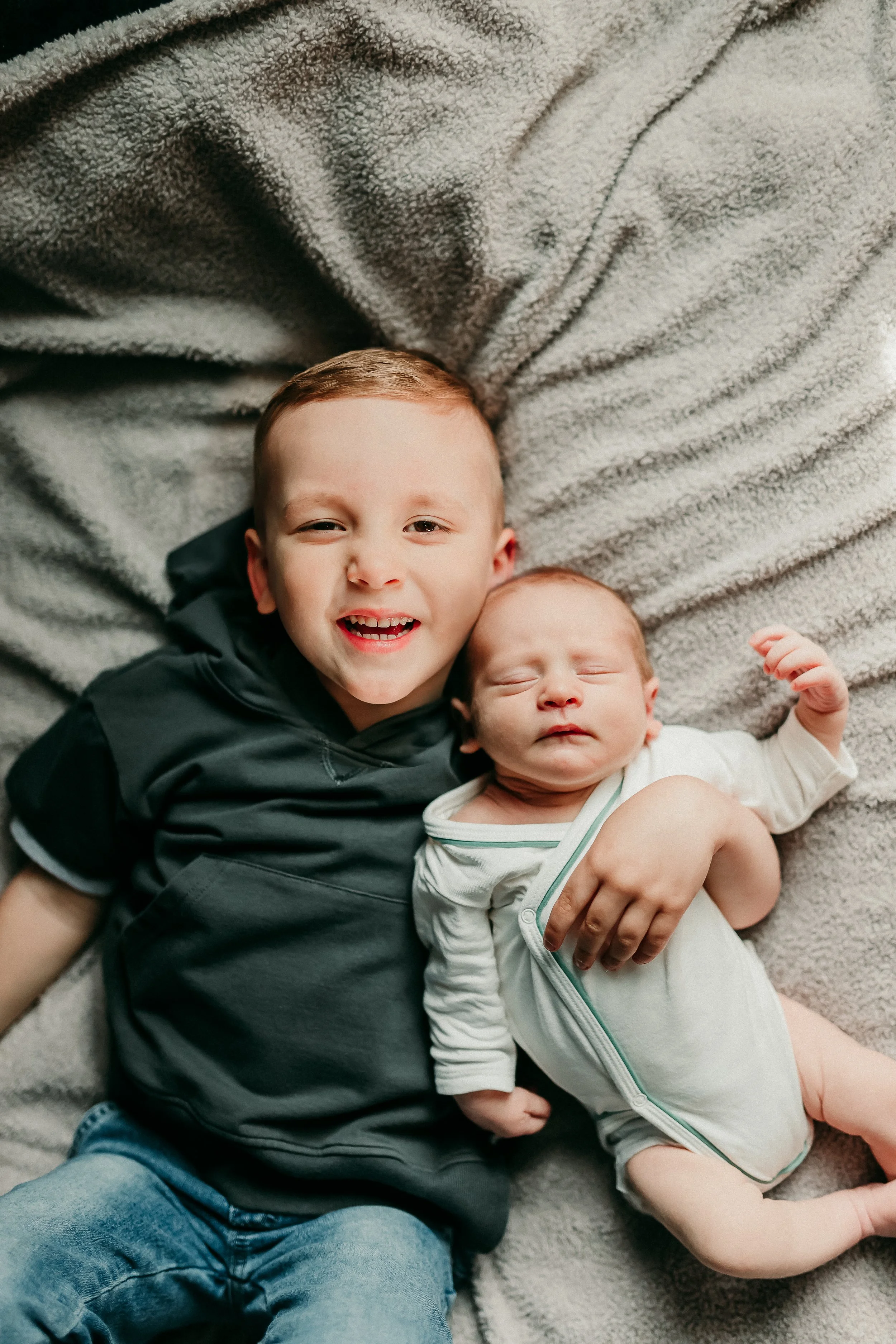 Two young children, a boy and a baby, lying on a soft, gray blanket. The older boy has short brown hair, is smiling, and is wearing a black hoodie and blue jeans. The baby has light hair, closed eyes, and is wearing a light-colored onesie.