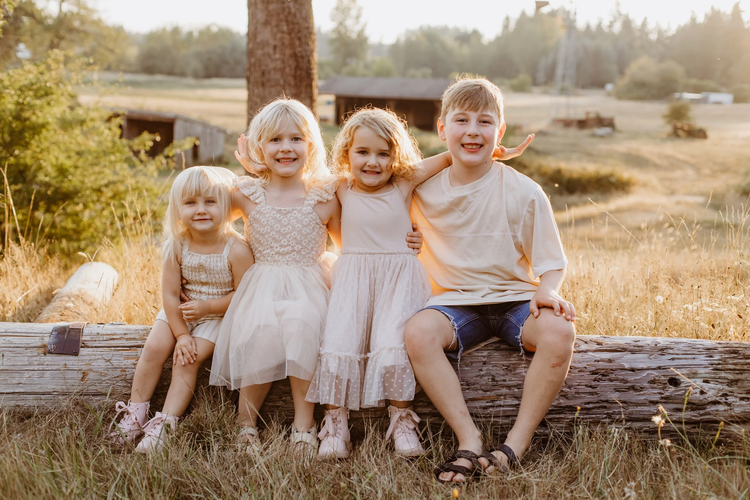 Four children sitting on a log outdoors in a field during sunset, smiling at the camera.