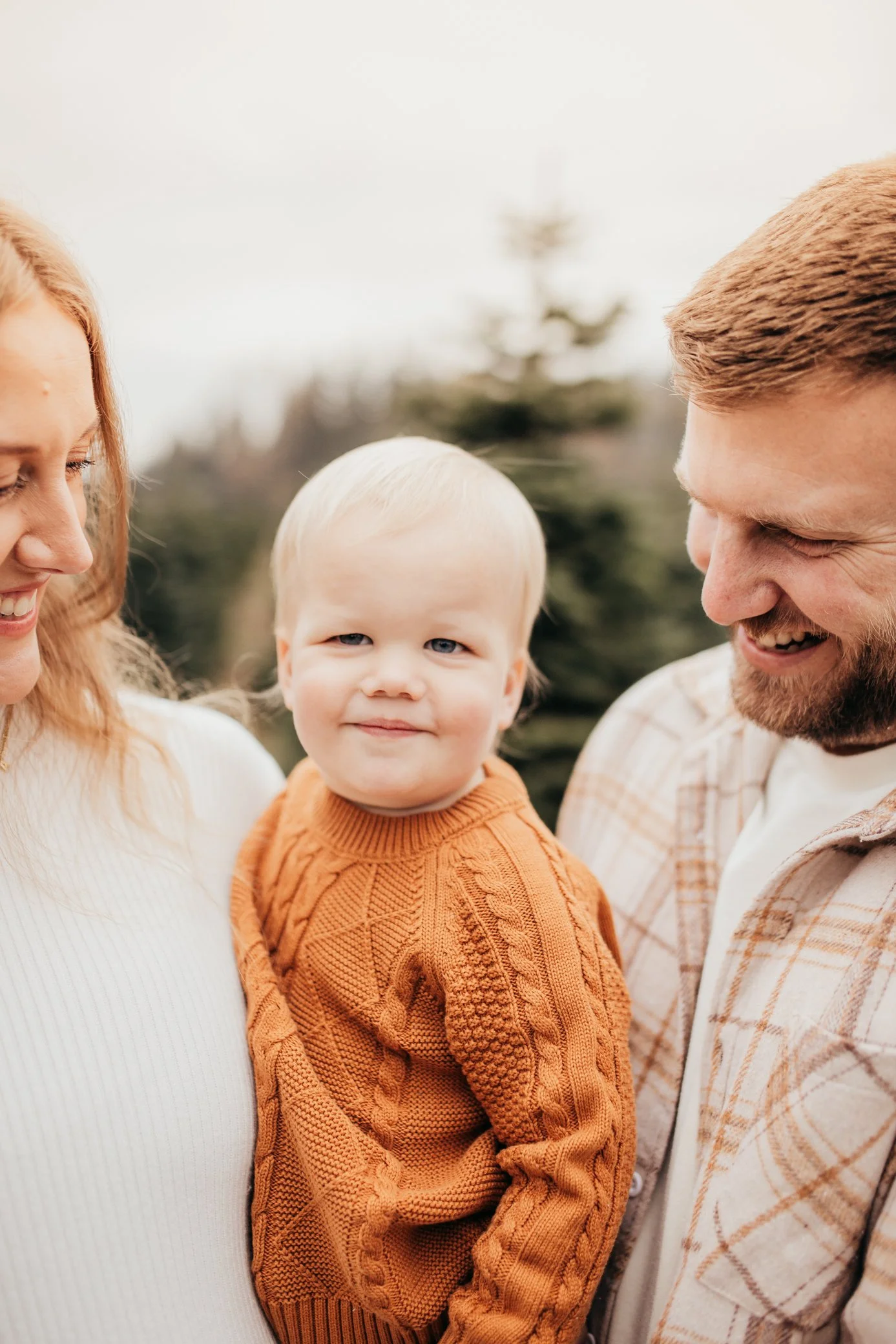 A smiling blonde toddler in an orange sweater is held by his mother on the left, while his father on the right also smiles. They are outdoors with a blurred background of trees.