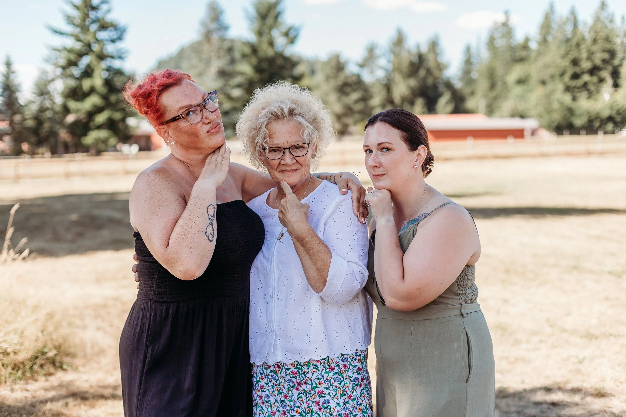Three women standing outdoors in a grassy area with trees in the background, posing for a photo. The woman in the middle is elderly with curly white hair, glasses, and wearing a white blouse and patterned skirt. The woman on the left has red hair, gl