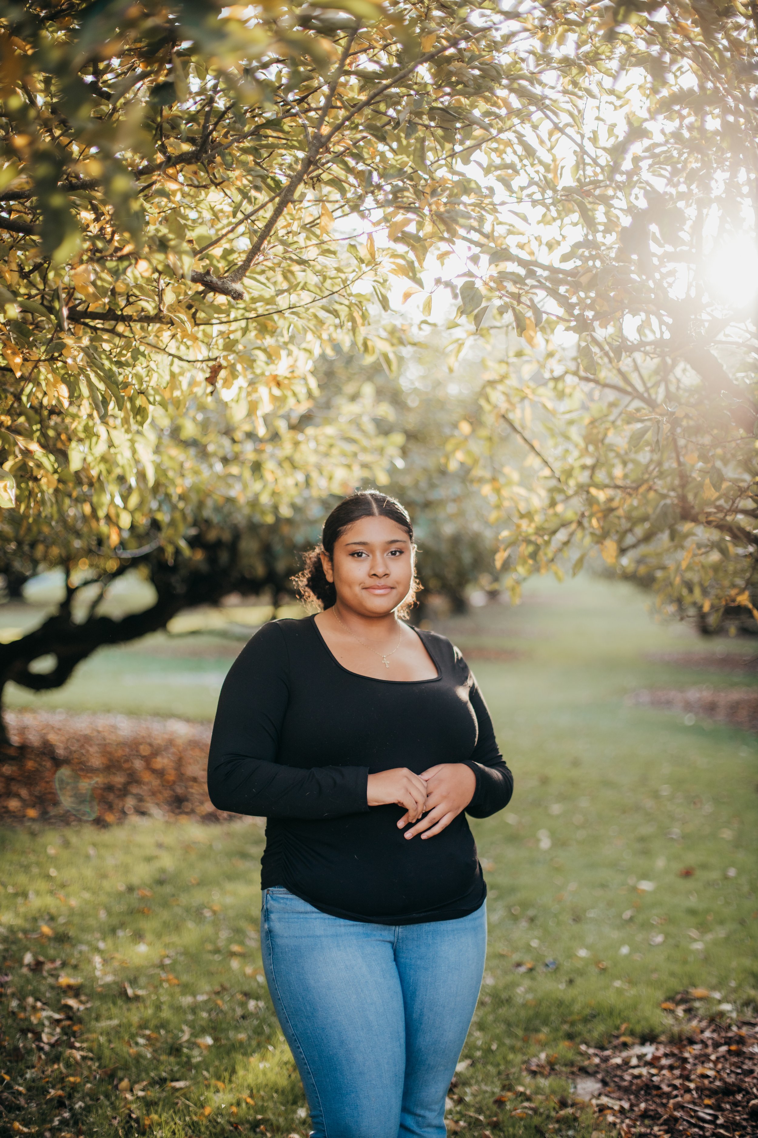 A woman with dark hair, wearing a black long-sleeve top and blue jeans, standing in a park with trees and sunlight filtering through the leaves.
