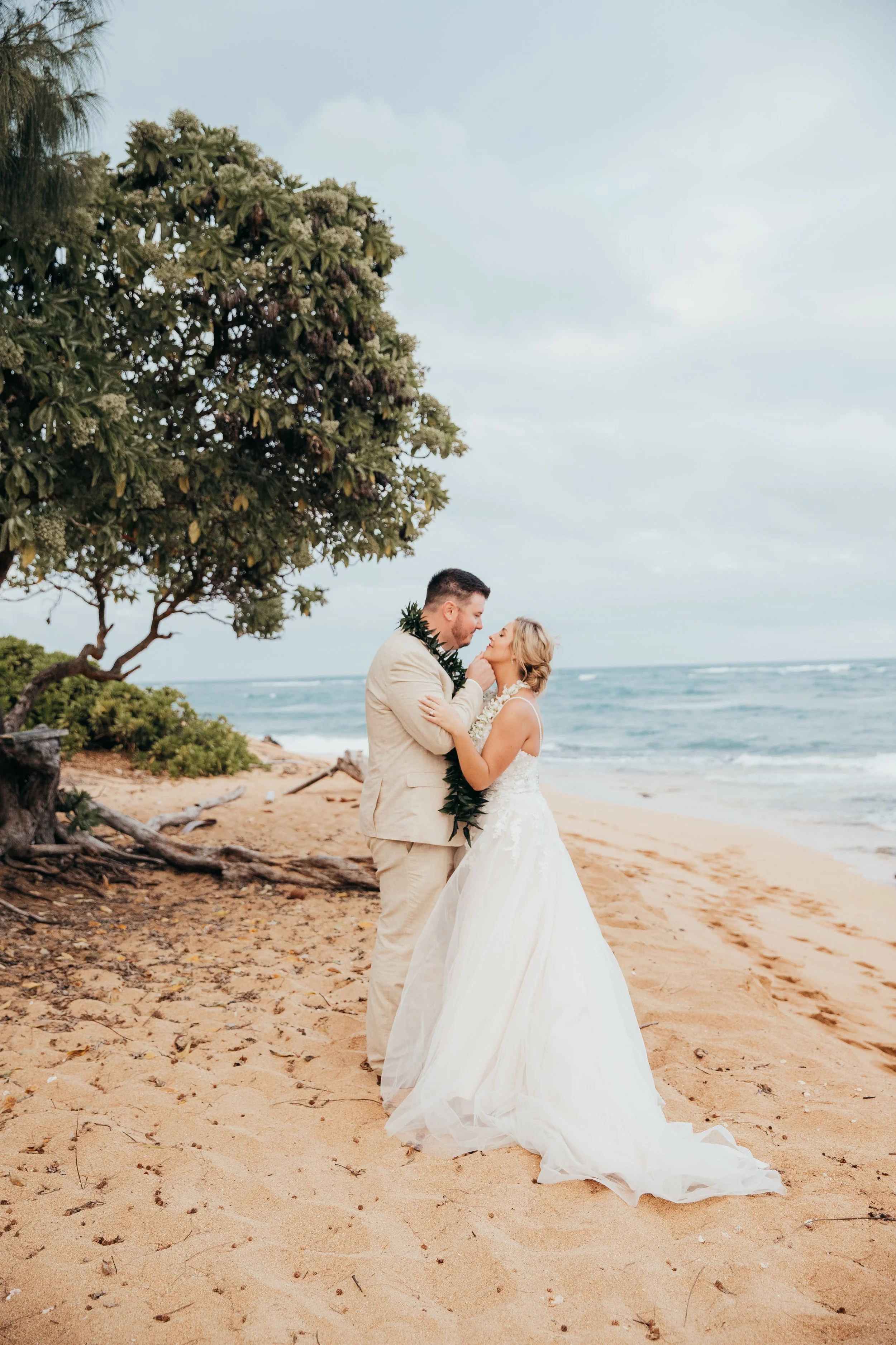 A bride and groom standing close together on a sandy beach, sharing an intimate moment with the ocean in the background. The bride is wearing a white wedding gown and the groom is in a beige suit, both adorned with a green leafy garland.