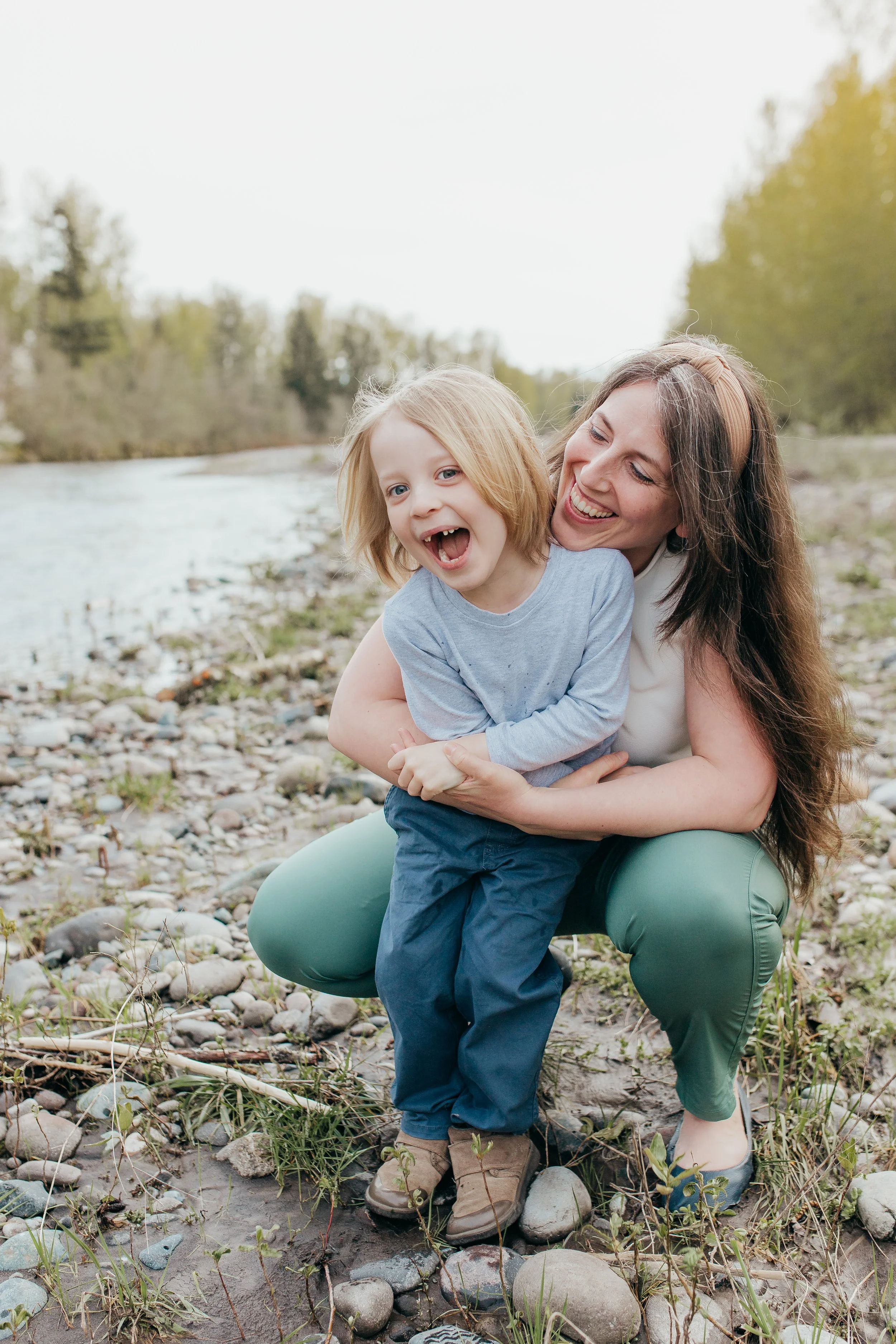 A woman and a young girl playing and laughing by a river on a rocky riverbank in a natural outdoor setting.