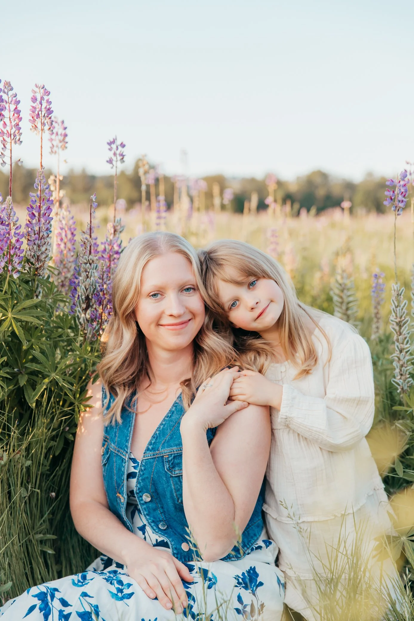 A woman and a young girl sitting in a field of tall purple and pink flowers, smiling and cuddling. The woman has blonde hair and is wearing a denim vest, while the girl has light brown hair and is wearing a light-colored dress.