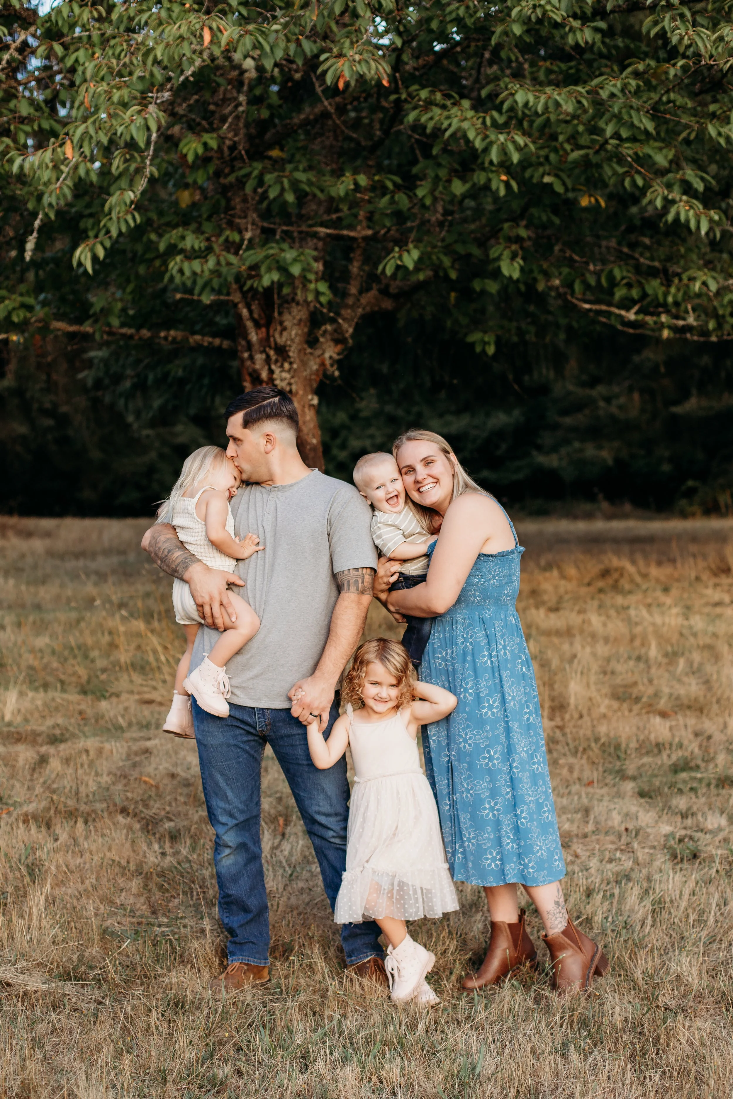 A family of five standing outdoors on grass with a large tree in the background. The father holds two young girls, and the mother holds a young boy, with a girl standing in front of the mother. Everyone is smiling and interacting fondly.