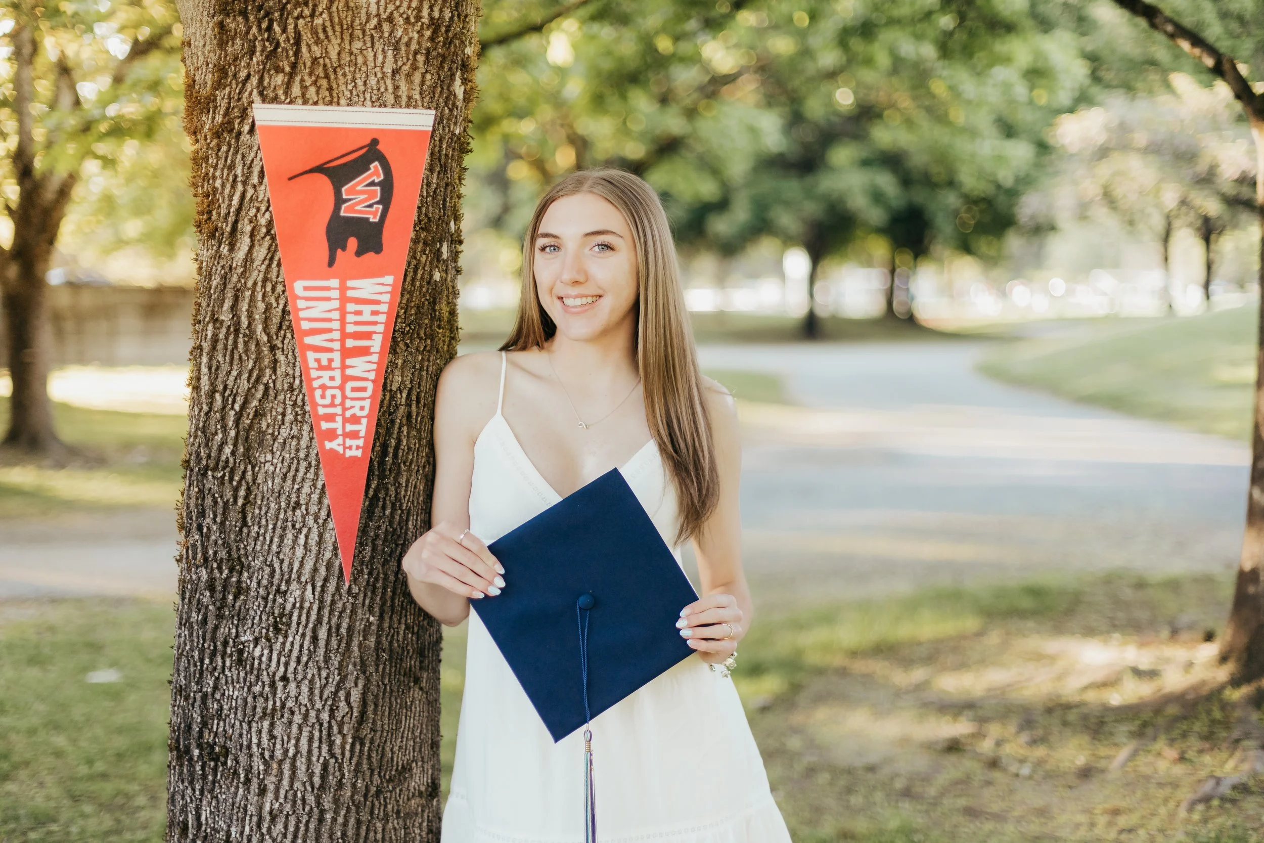 A young woman in a white dress holding a graduation cap standing outdoors next to a tree with a maroon and white university pennant hanging on it, in a park-like setting with trees and a pathway in the background.