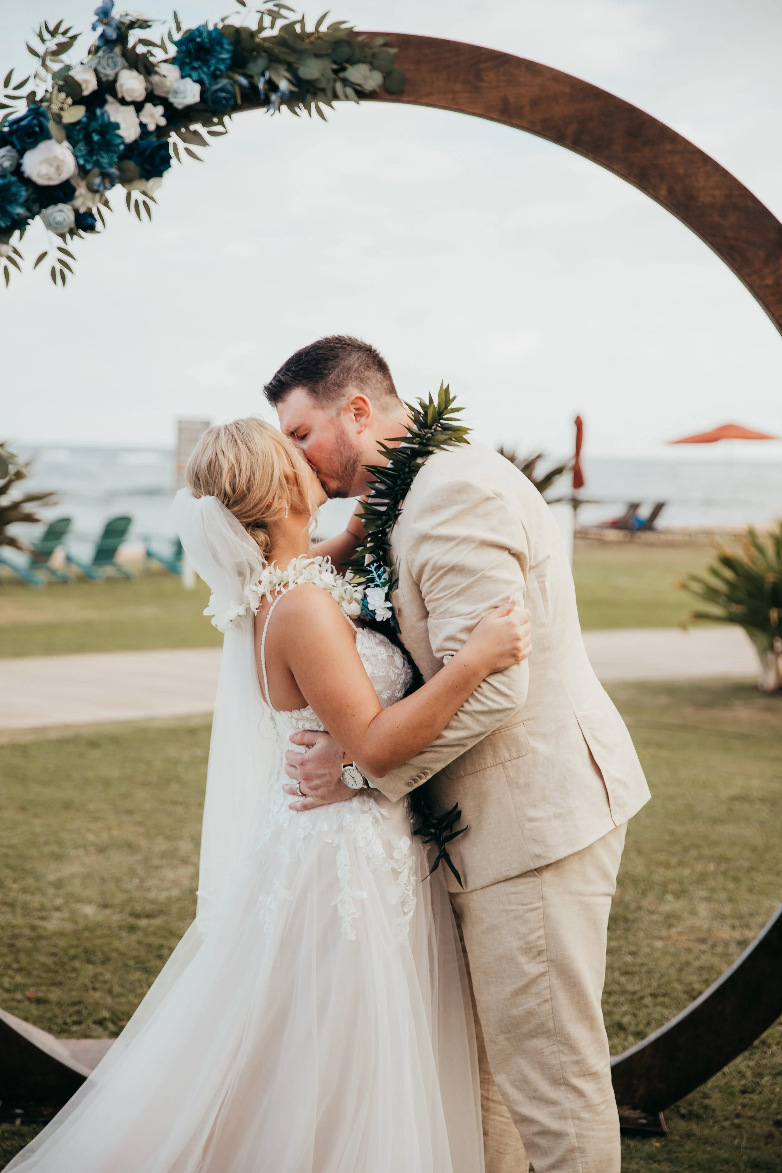 A couple kissing during a wedding ceremony on the beach, with the ocean and beach chairs in the background.