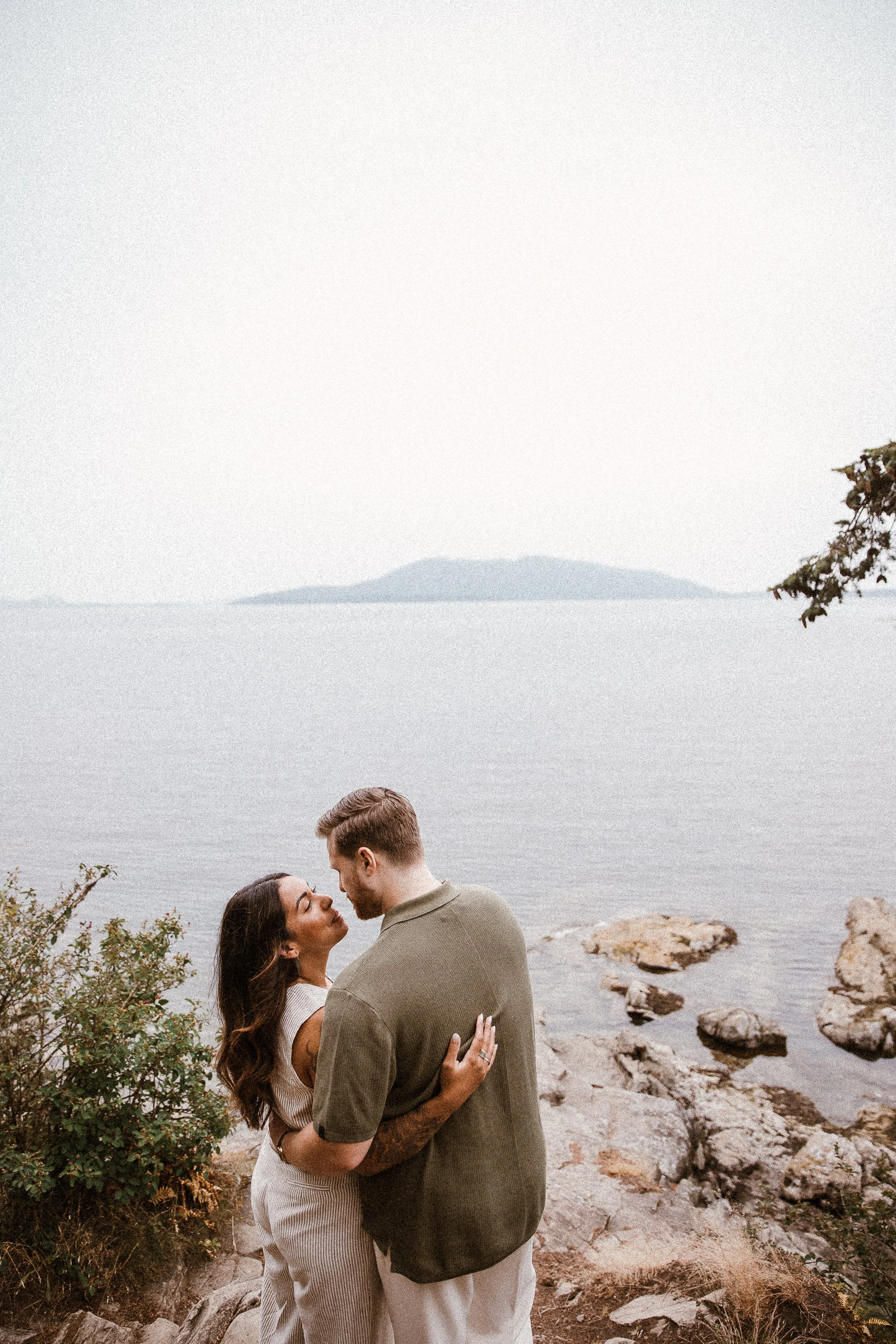 A couple standing close to each other on a rocky shoreline near a body of water, with an island in the distance.