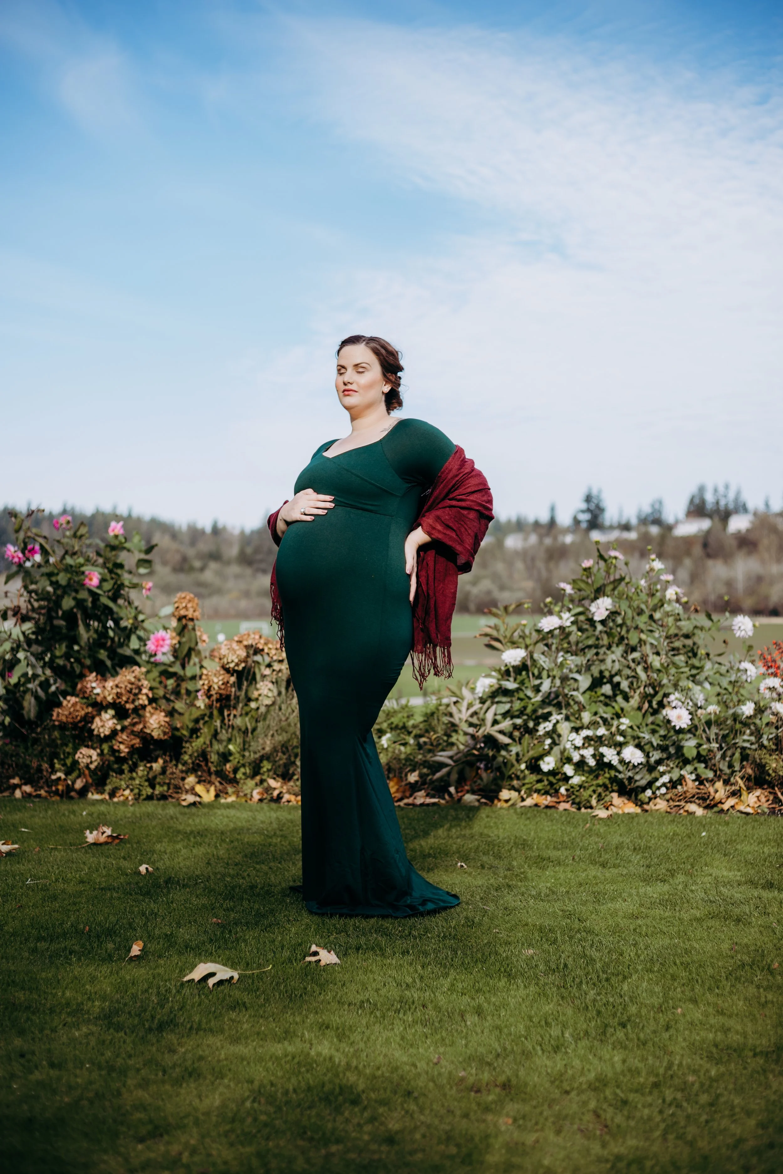 A pregnant woman in a long green dress standing outdoors on a grassy area with flowering bushes and trees in the background under a partly cloudy sky.