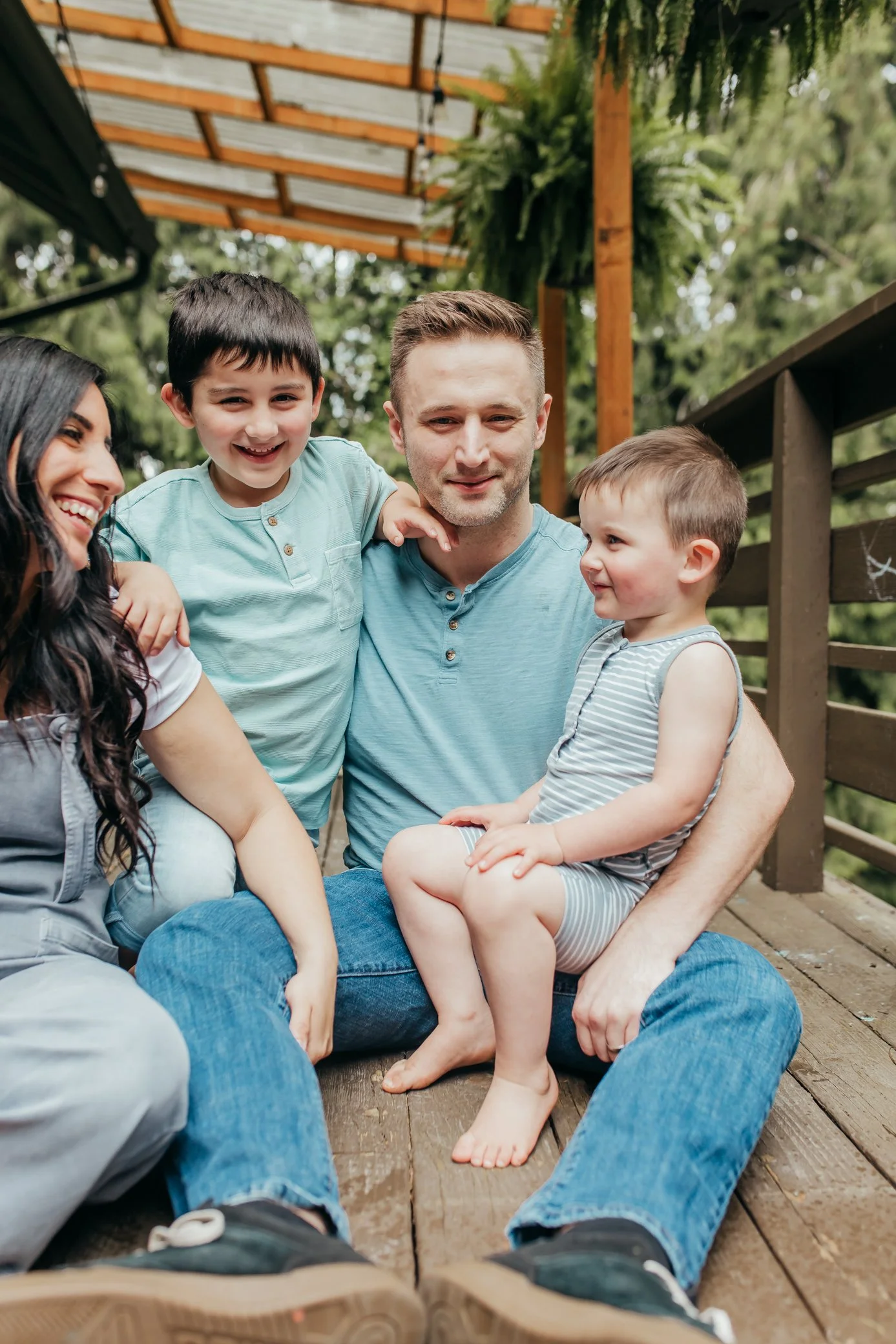 A family of four sitting on a wooden porch outdoors, smiling and interacting. The father is holding a young boy, and they are joined by a woman and another young boy, all enjoying a happy moment in a lush green background.