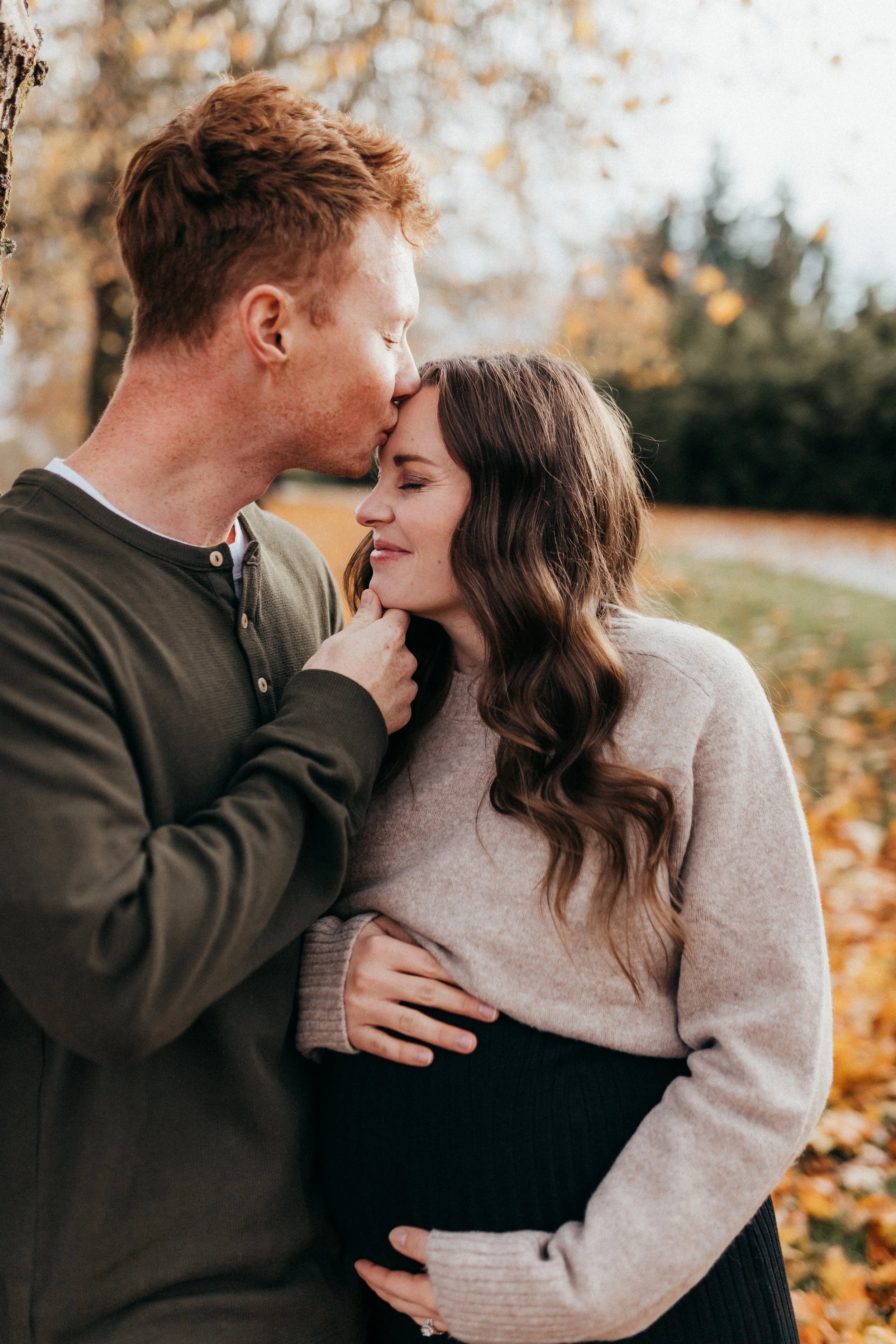 A man kisses a pregnant woman on the forehead while she smiles with her hand on her belly outdoors during fall.