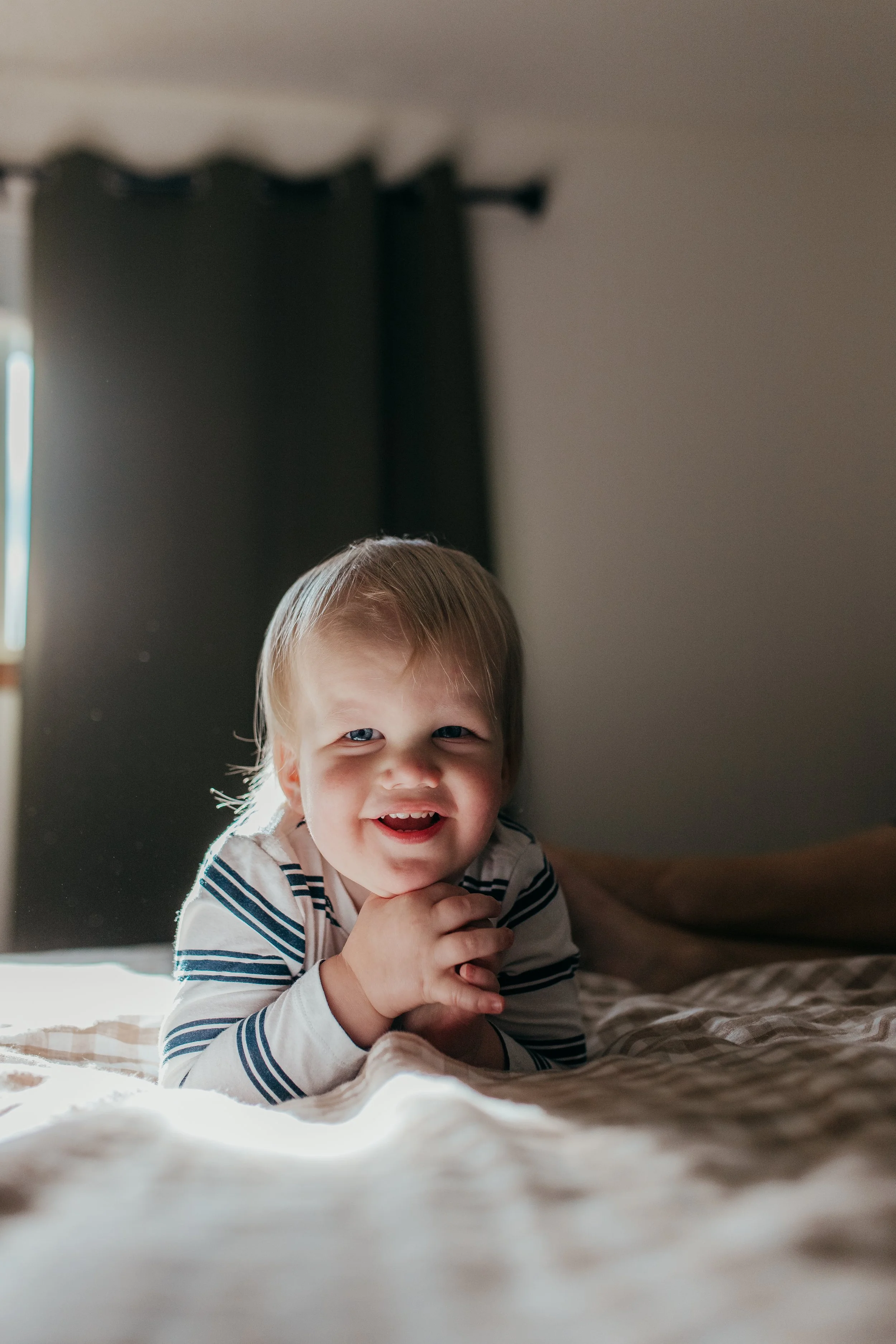 A happy young boy with blonde hair and blue eyes, smiling and lying on a bed with striped pajamas, with sunlight coming through a dark curtain in the background.