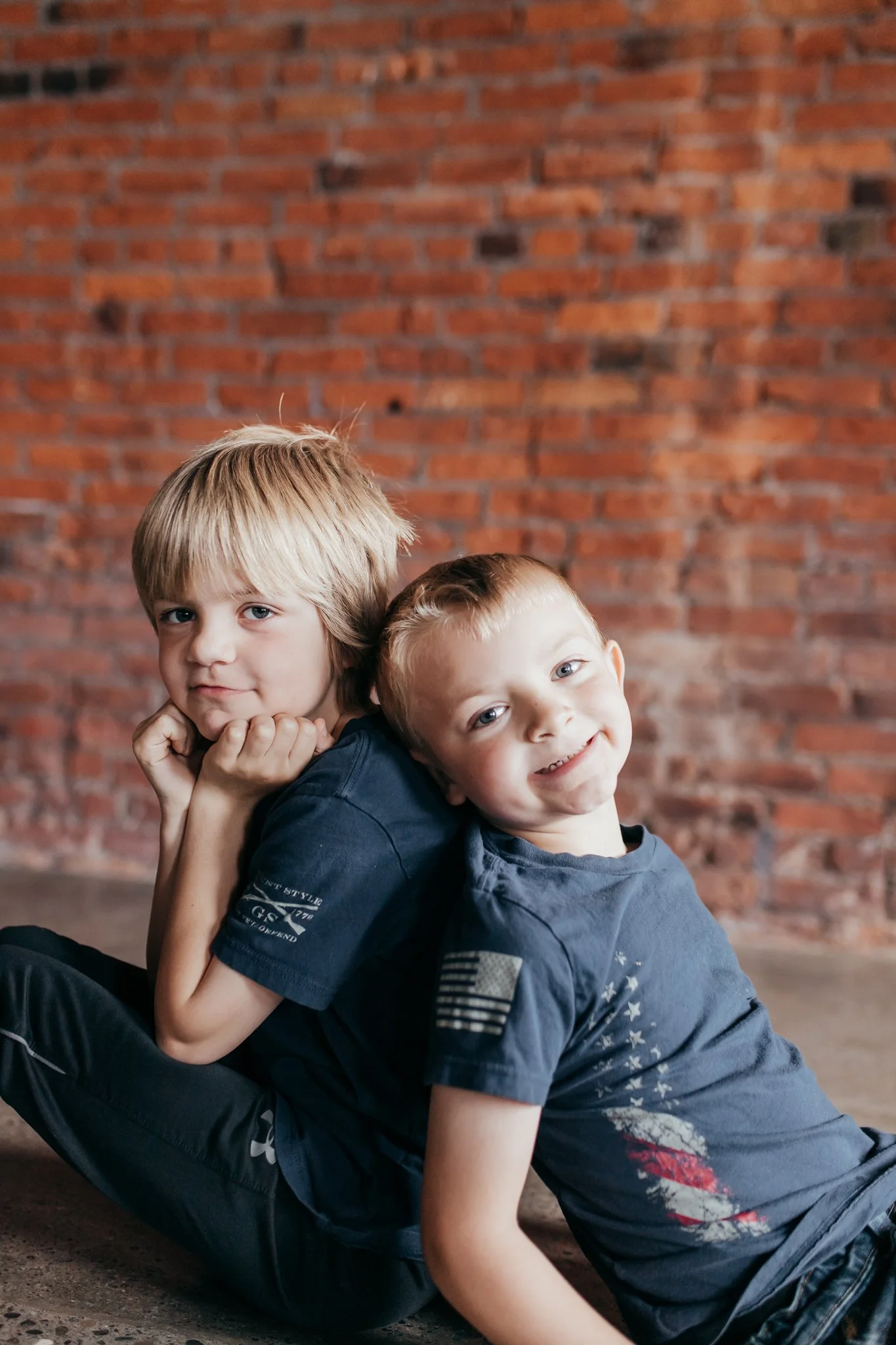 Two young boys sitting back-to-back on the floor in front of a brick wall, with one smiling and the other resting his chin on his hand.
