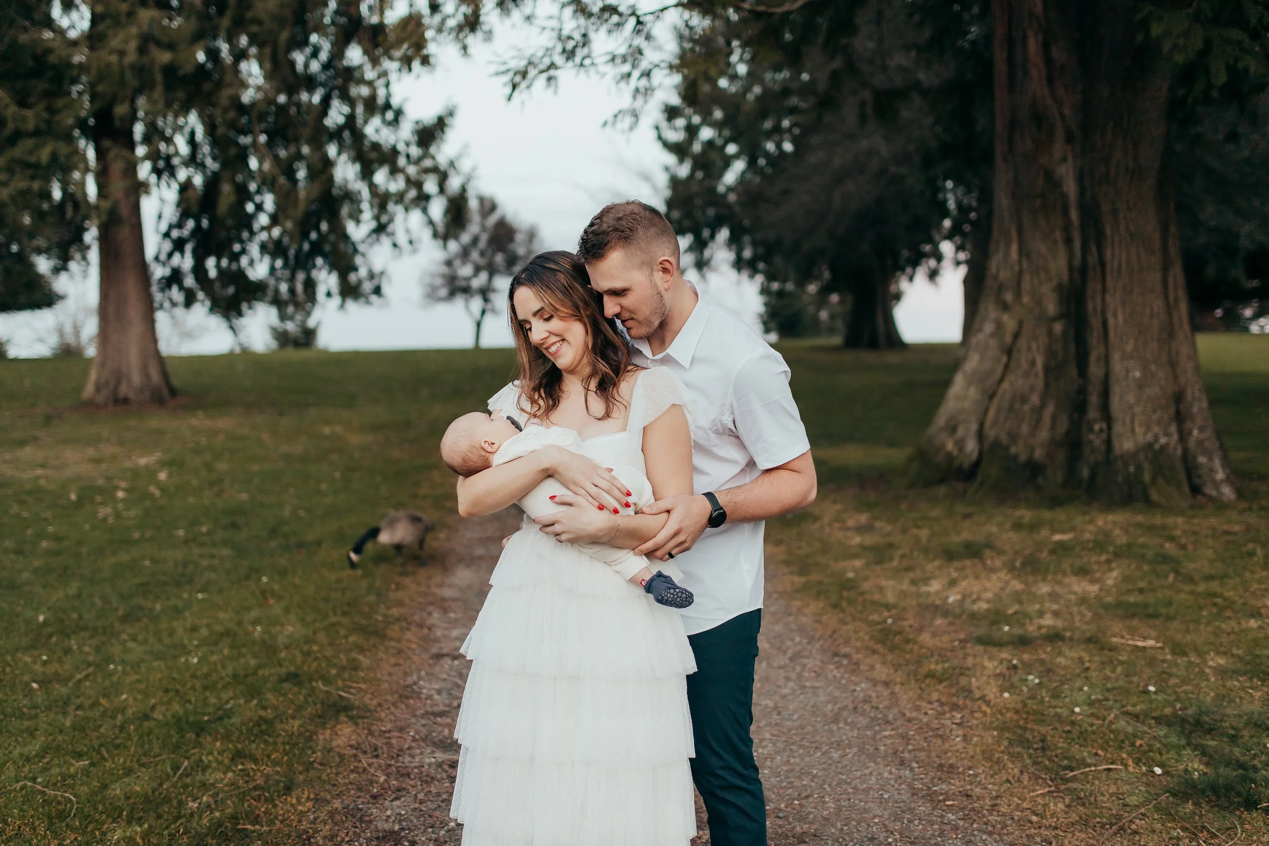 A couple holding a young child outdoors in a park with large trees in the background.
