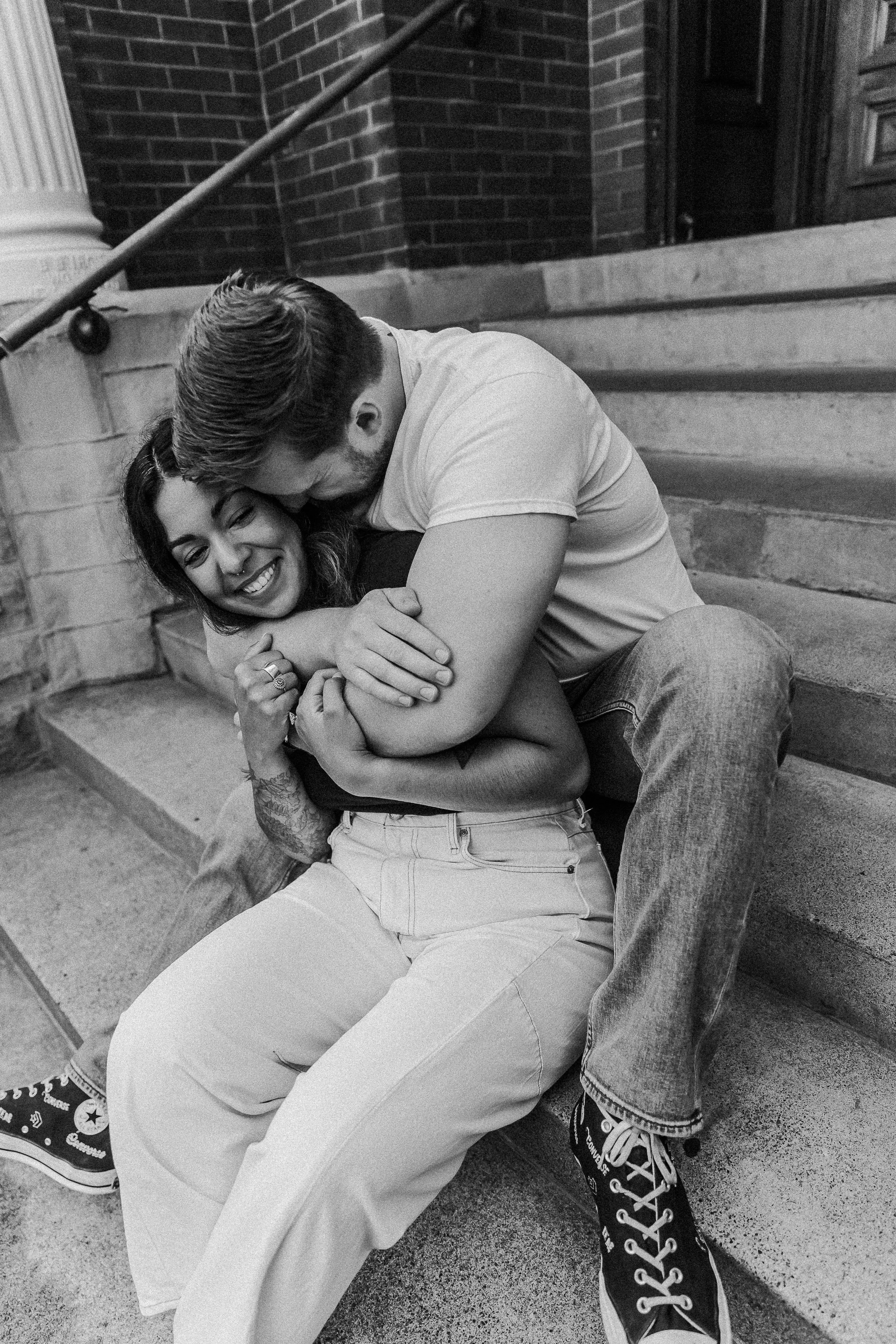 A black-and-white photo of a couple sitting on steps outside, embracing and smiling, with the woman looking happy and the man kissing her on the head.
