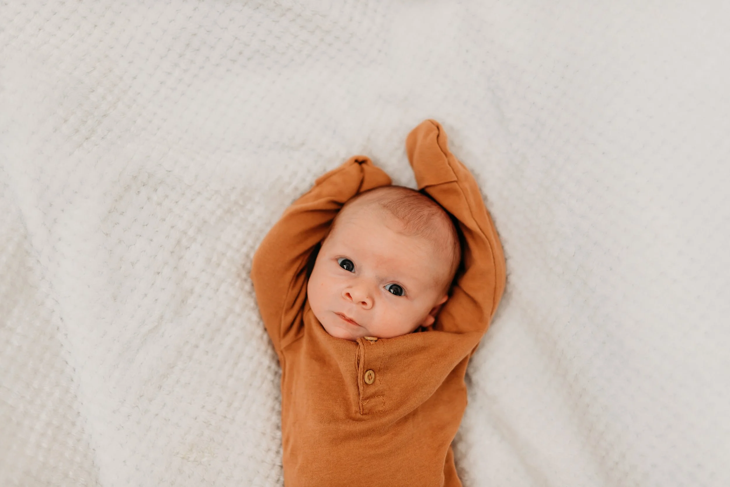 A baby lying on a textured white blanket, wearing a brown onesie with their arms raised above their head, looking directly at the camera.