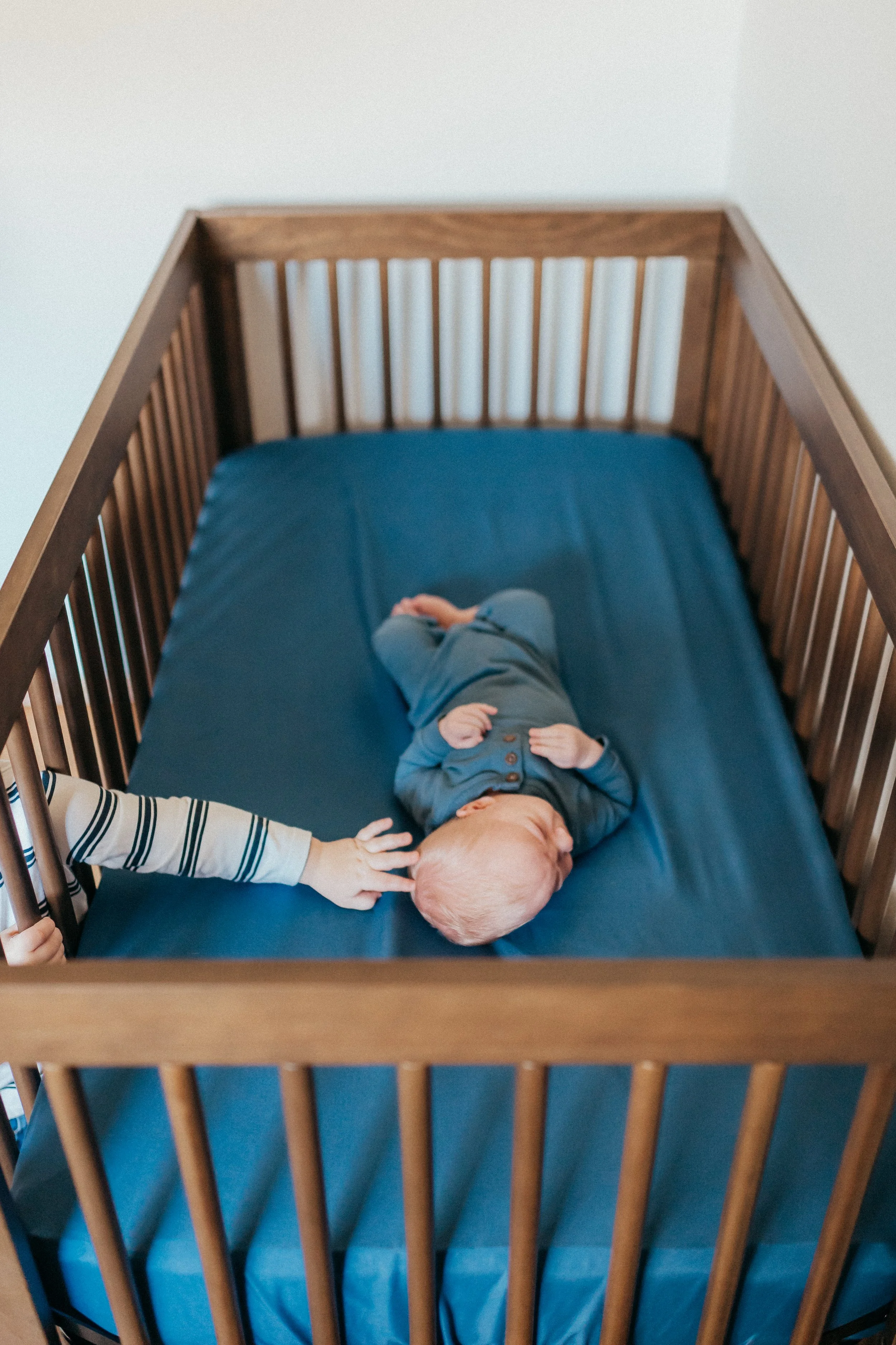 A baby lies on a blue mattress inside a wooden crib, with a child's hand reaching out to touch the baby's head.
