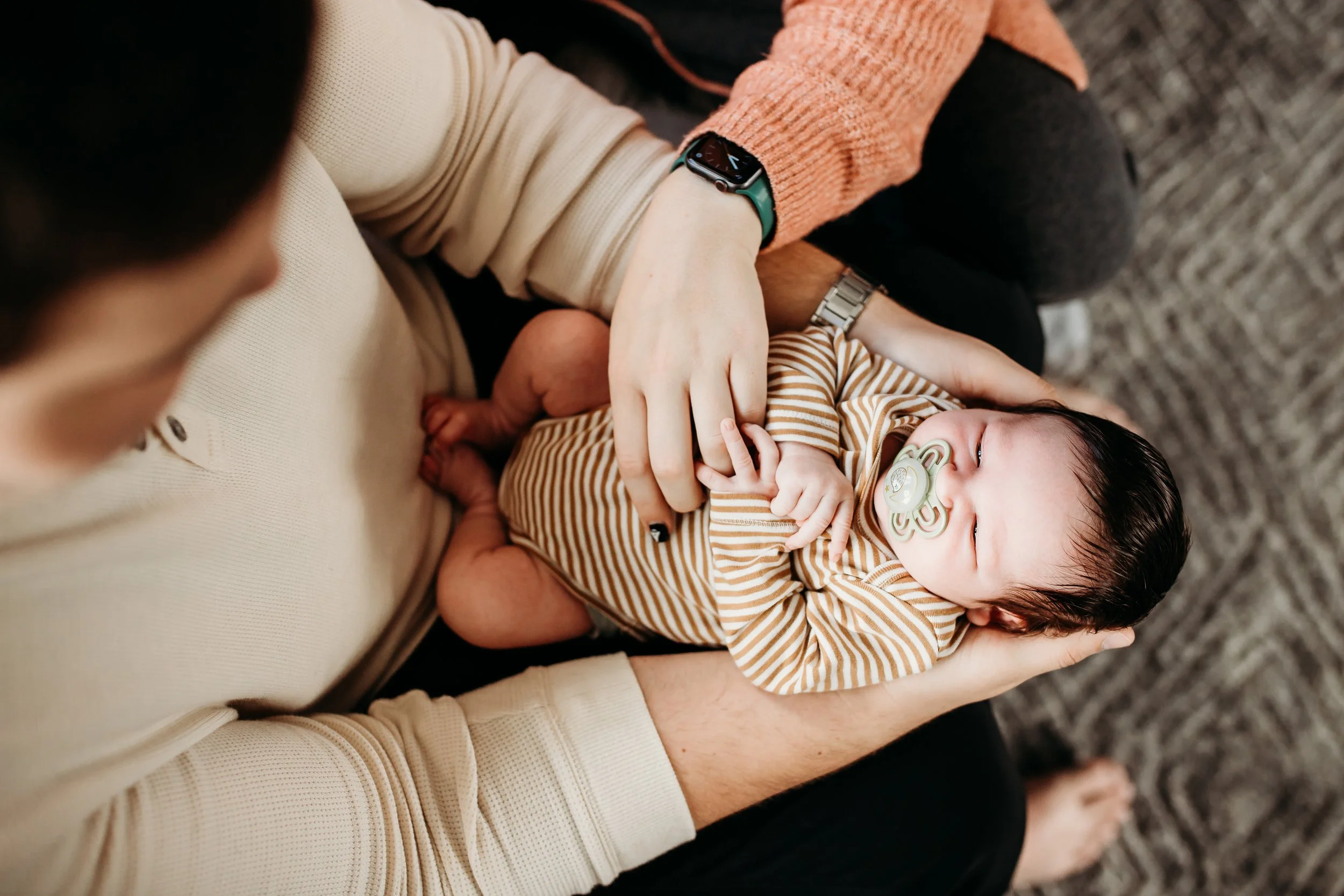 A person holding a sleeping baby with a pacifier, sitting on a gray carpet.