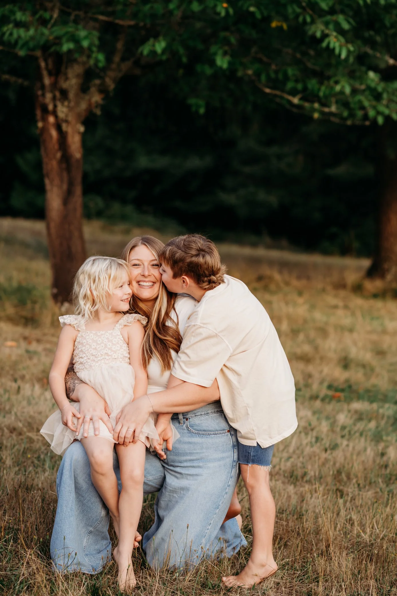 A woman is kneeling on grass with two children, a girl and a boy, in a wooded outdoor setting. The girl is sitting on the woman's lap, and the boy is kissing her cheek. The scene is warm and joyful.