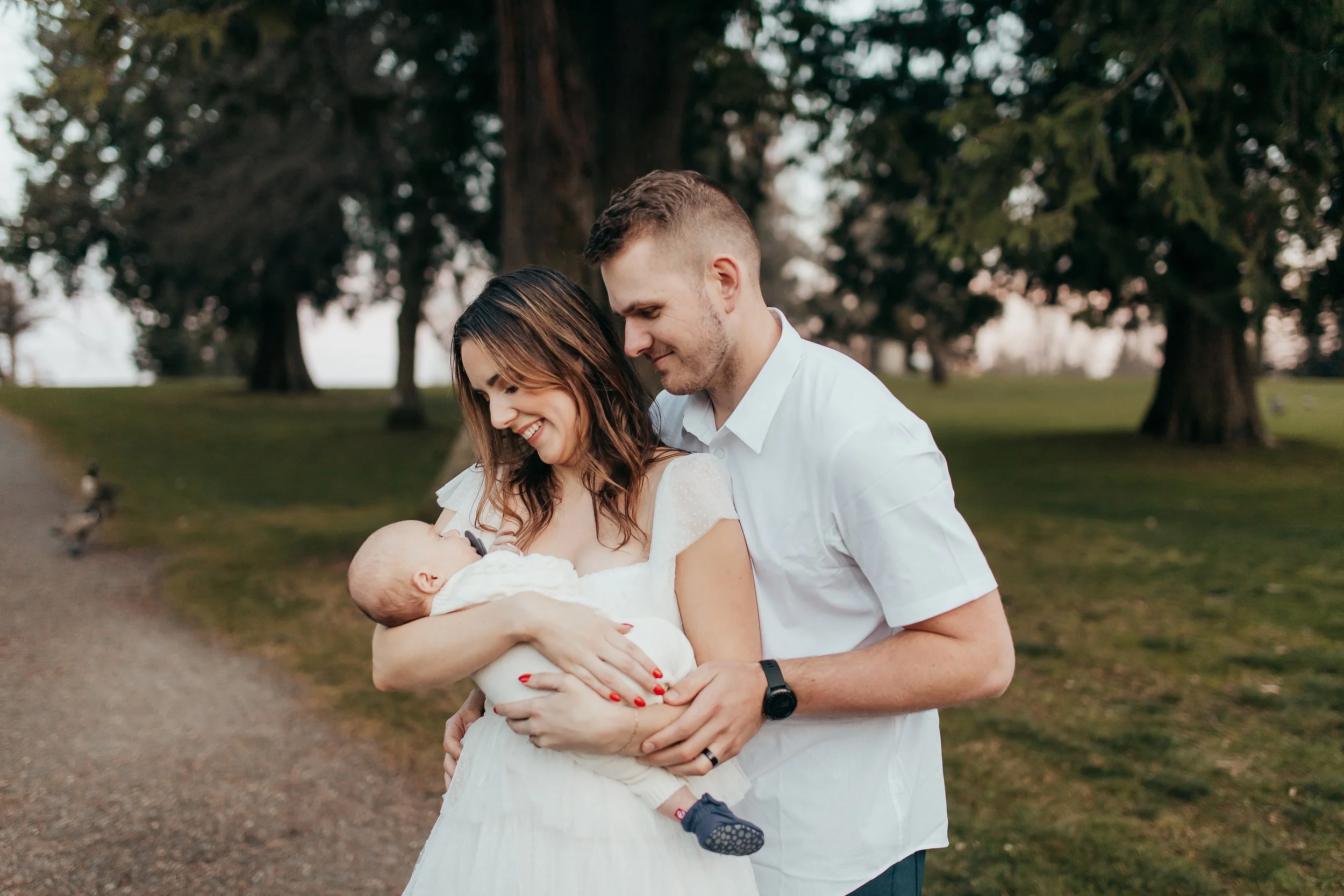 A happy family in a park with large trees, a woman holding a baby and a man standing beside her, all smiling and looking at the baby.