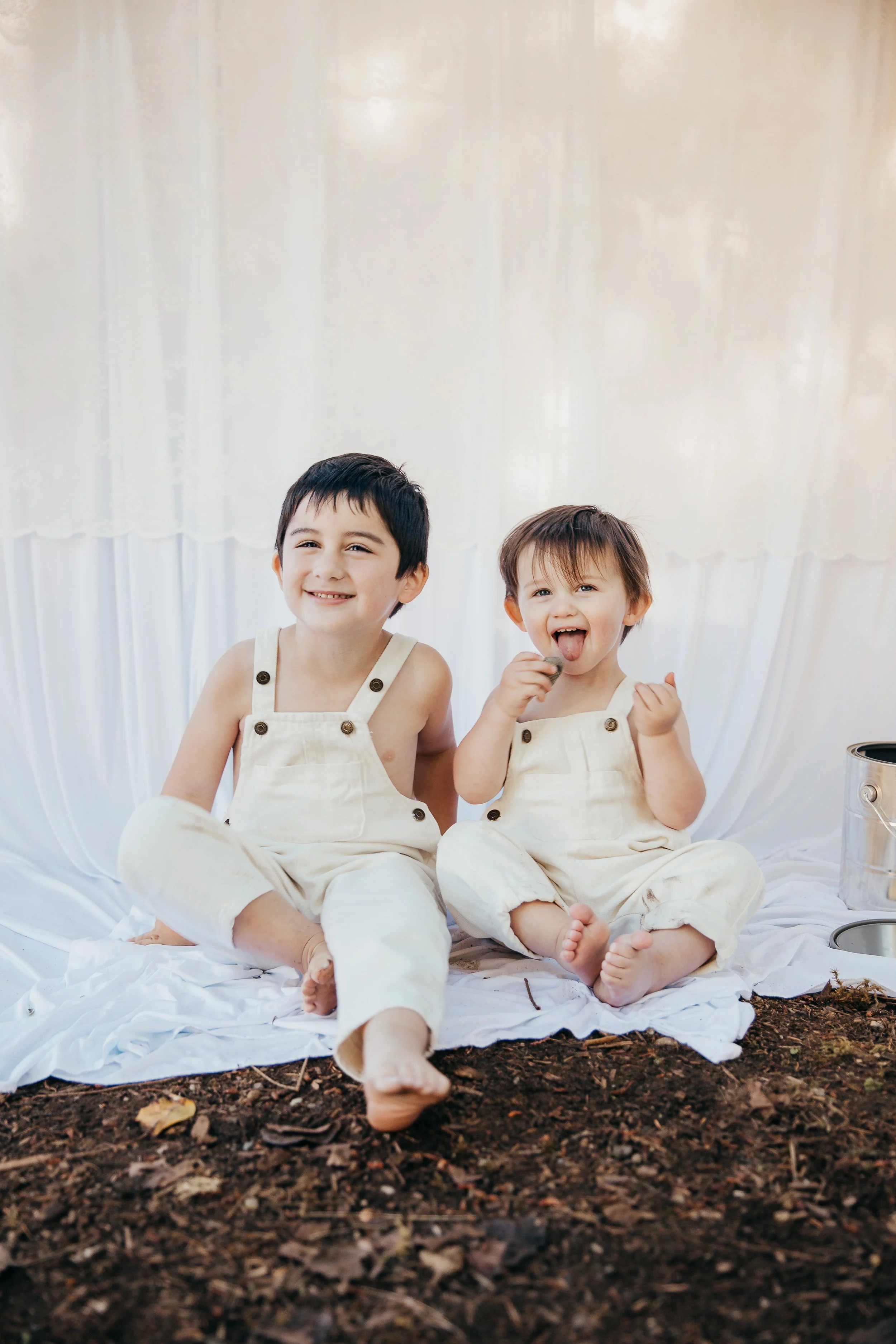 Two young boys sitting on white cloth in front of a white backdrop, with gardening tools and soil in front, dressed in off-white overalls, smiling and playing.