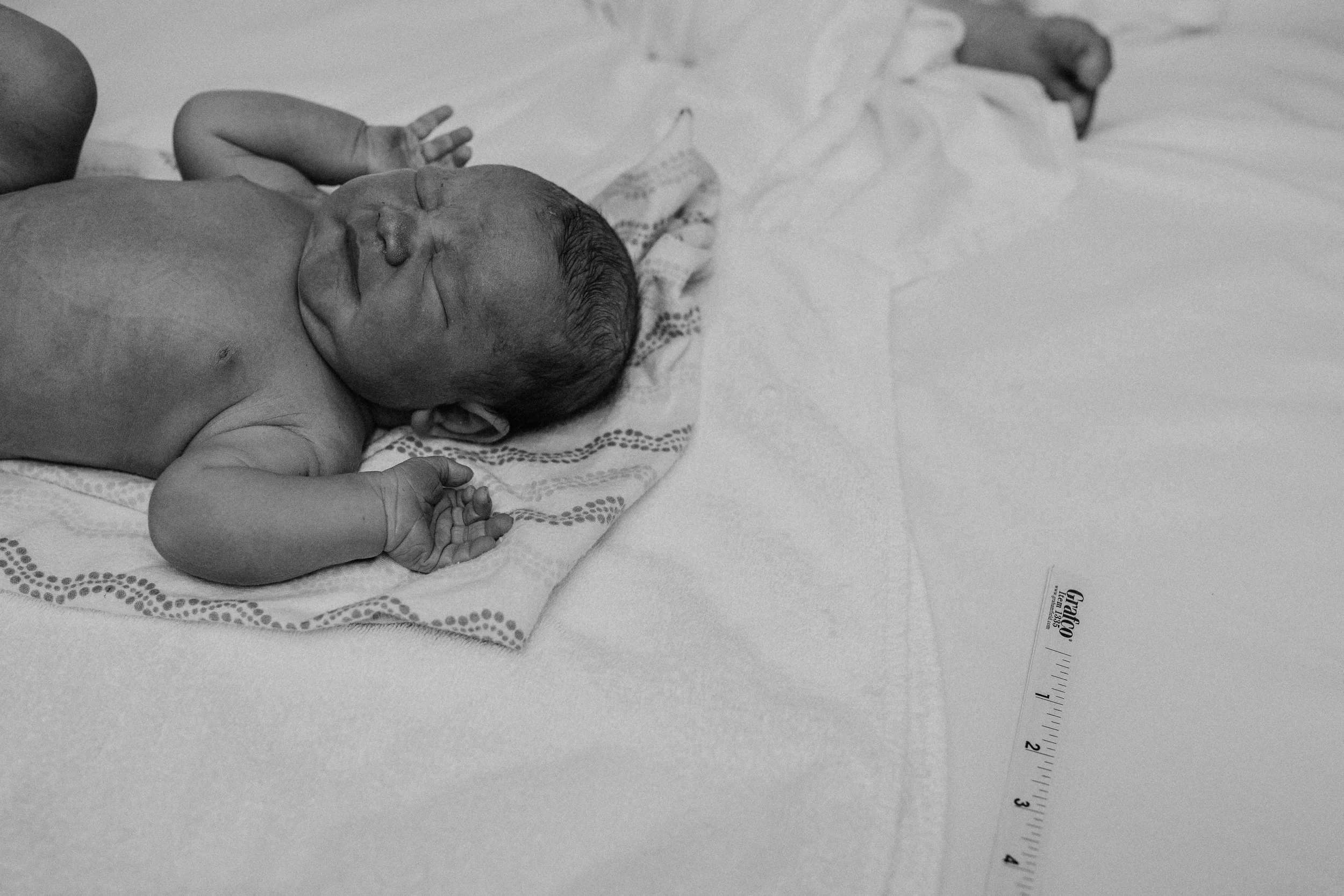 A newborn baby lying on a patterned cloth on a bed with a ruler beside, in black and white.