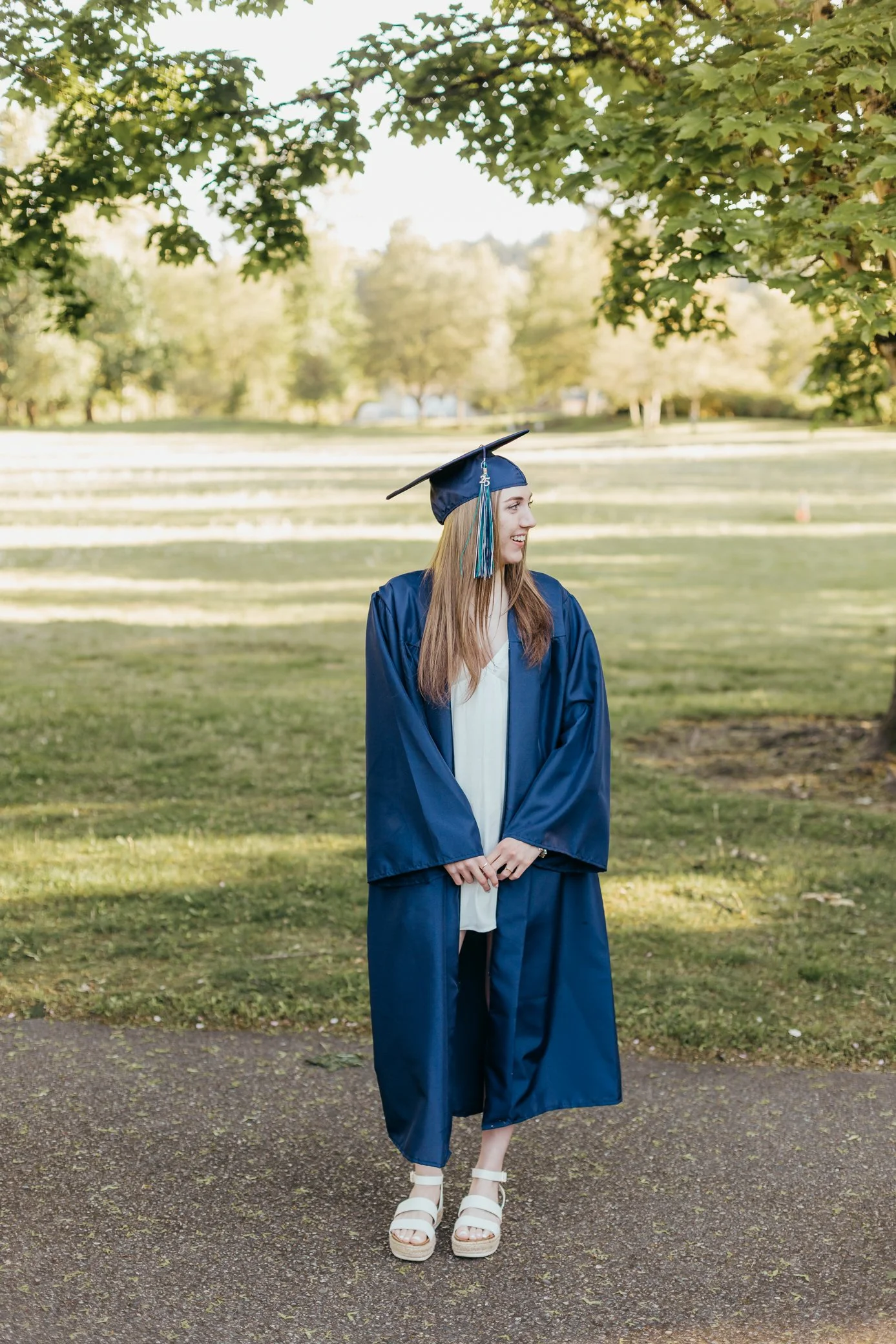 A young woman wearing a blue graduation gown and cap with blue tassel stands on a pathway outdoors, smiling and looking to her left, with trees and an open grassy area in the background.