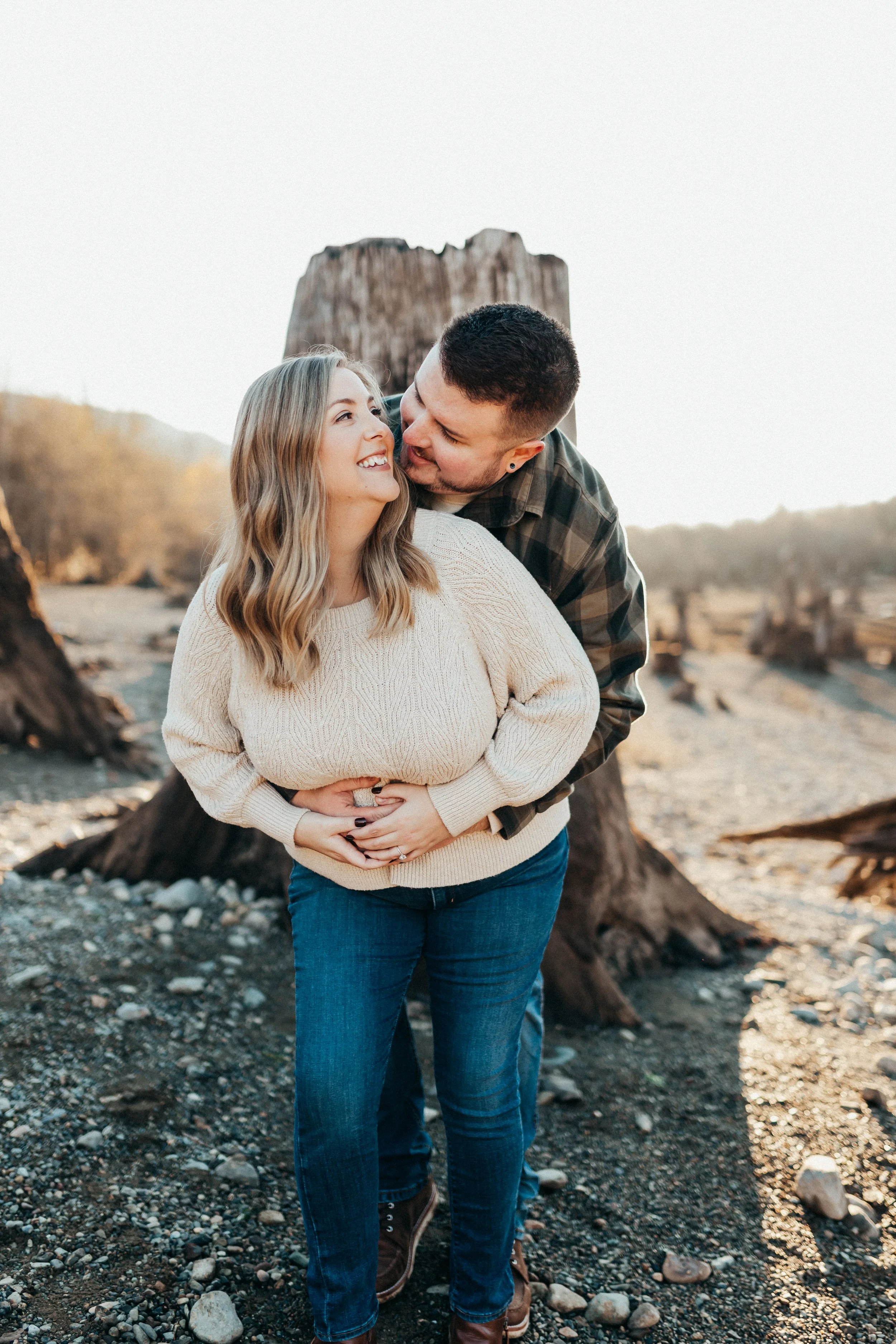 a smiling couple embracing outdoors near a large tree trunk, with rocky ground and a distant natural landscape in the background