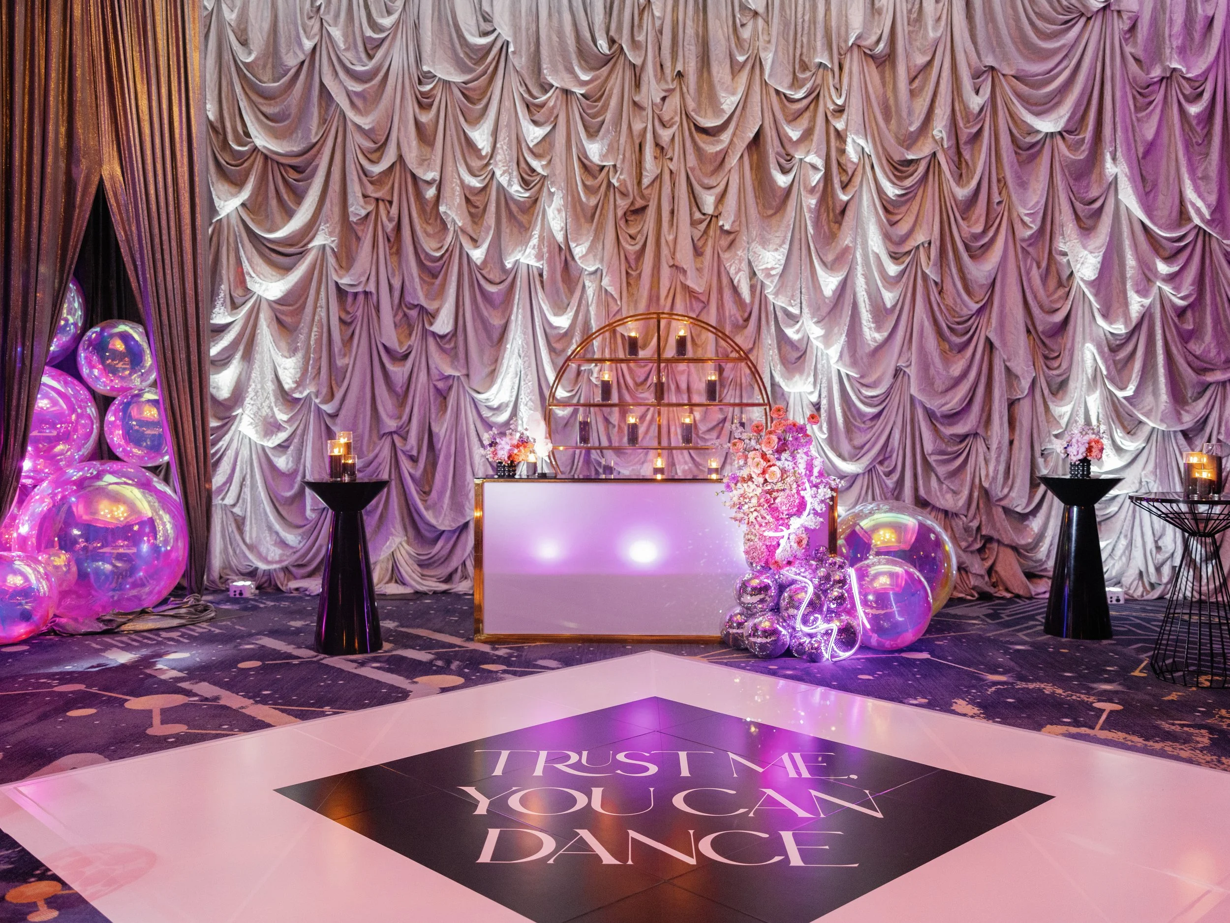 Decorative party setup with a dance floor that says 'Trust me, you can dance,' surrounded by black tables, candles, pink flowers, iridescent balloons, and draped pink curtains.