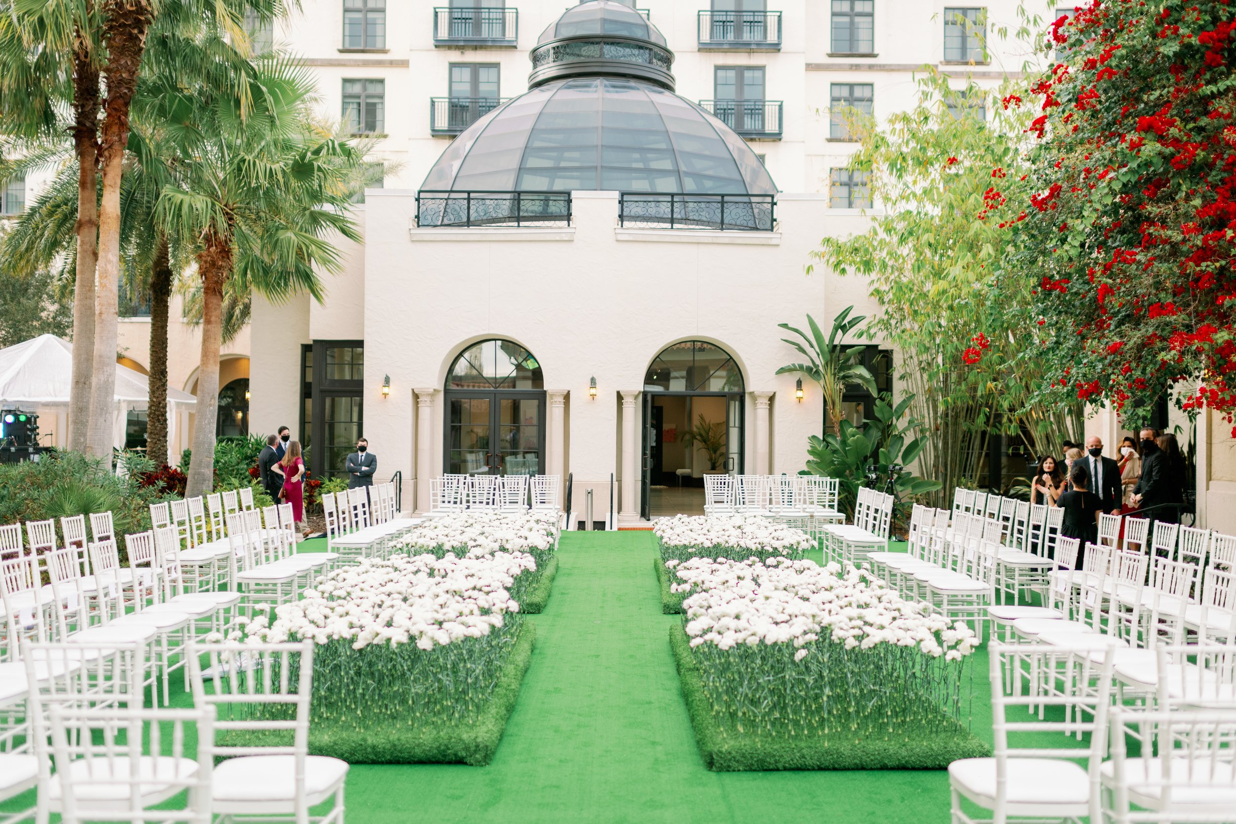 A wedding ceremony setup outdoors with white chairs arranged on either side of a green aisle, white flowers lining the aisle, and a white building with a glass dome in the background, surrounded by palm trees and greenery.