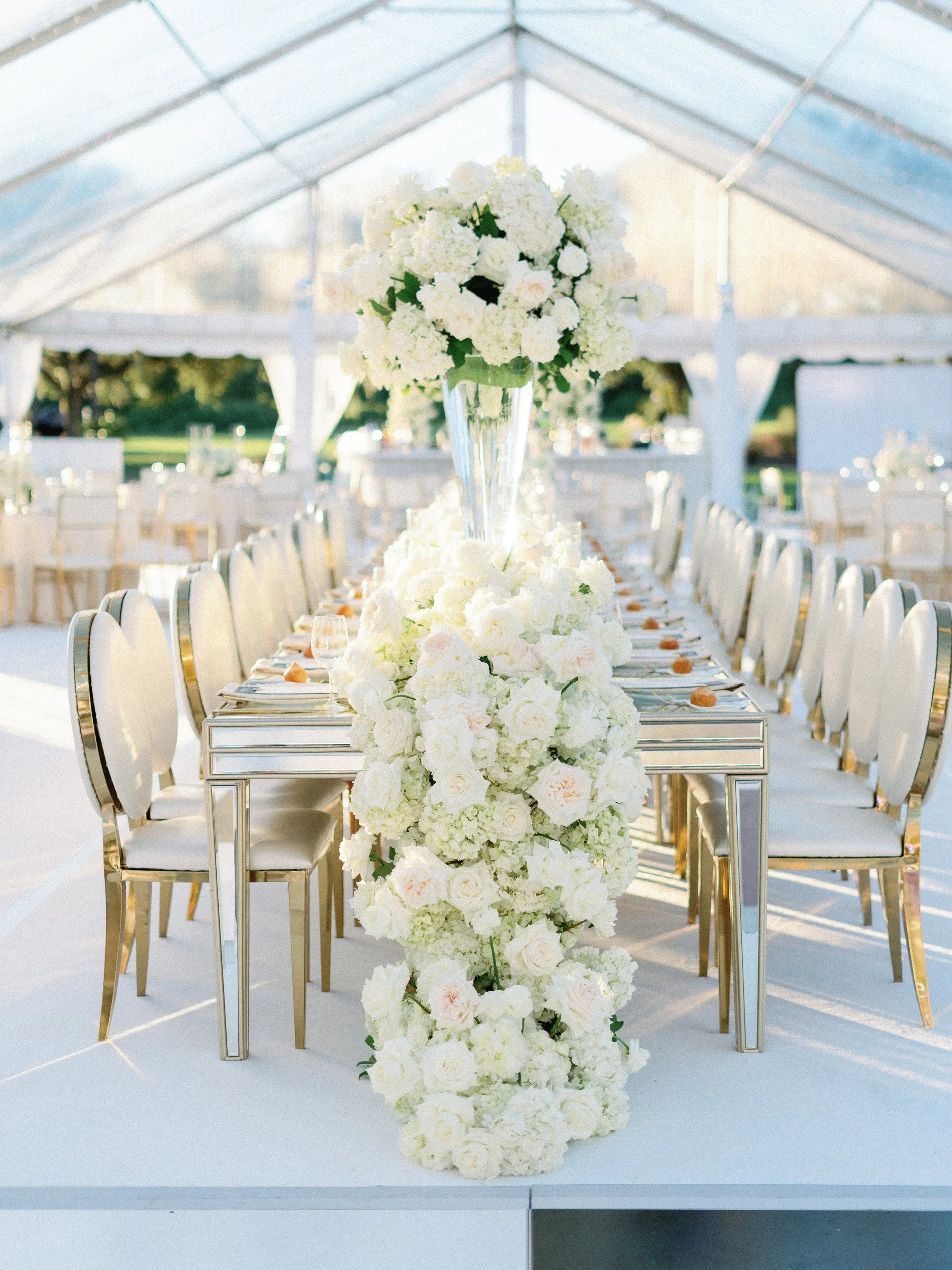 Elegant wedding reception setup with a table decorated with white flowers and a tall floral centerpiece in a glass vase under a glass tent.