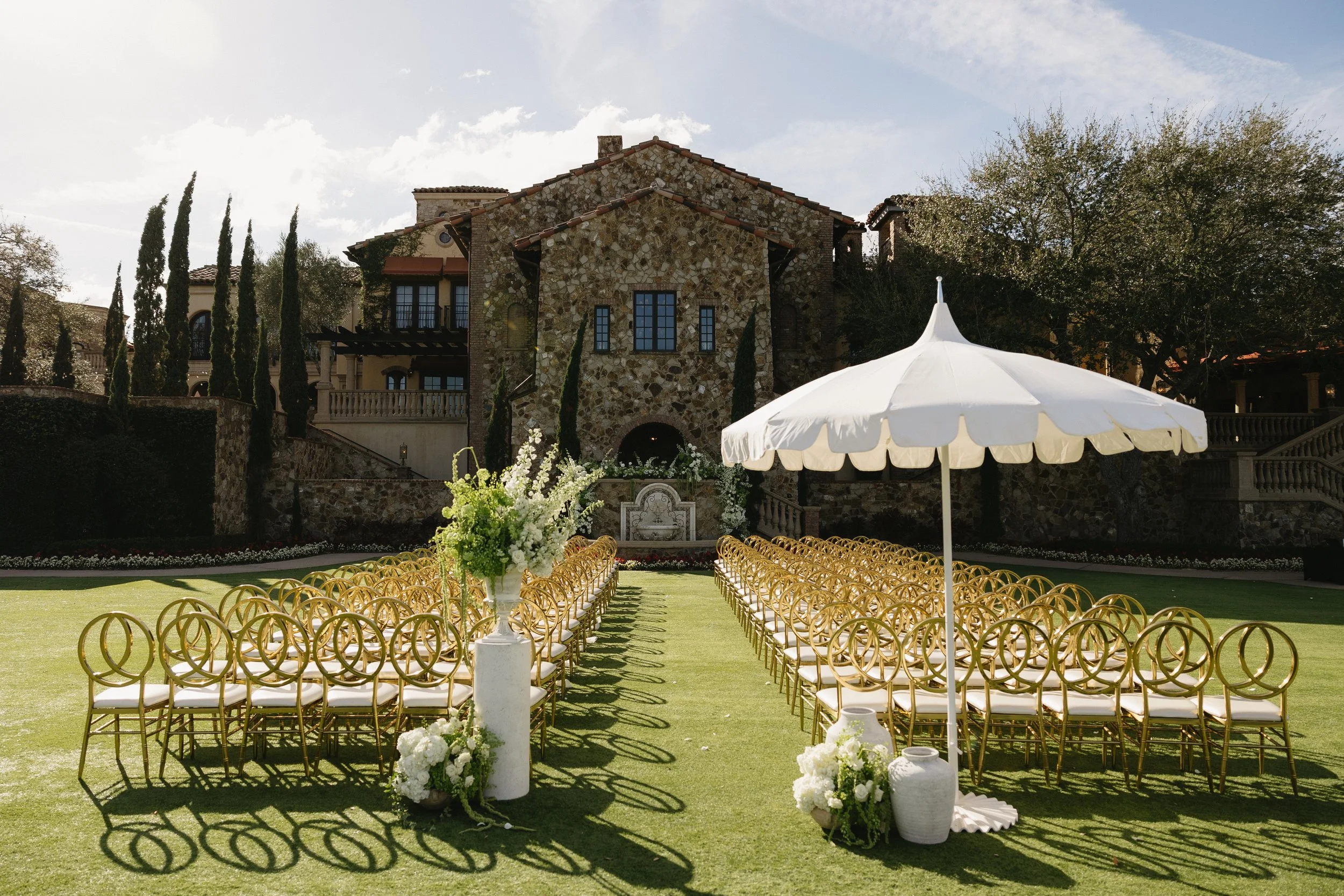 Outdoor wedding setup with gold chairs arranged in rows, white floral arrangements, a white parasol, and a large stone house in the background on a sunny day.