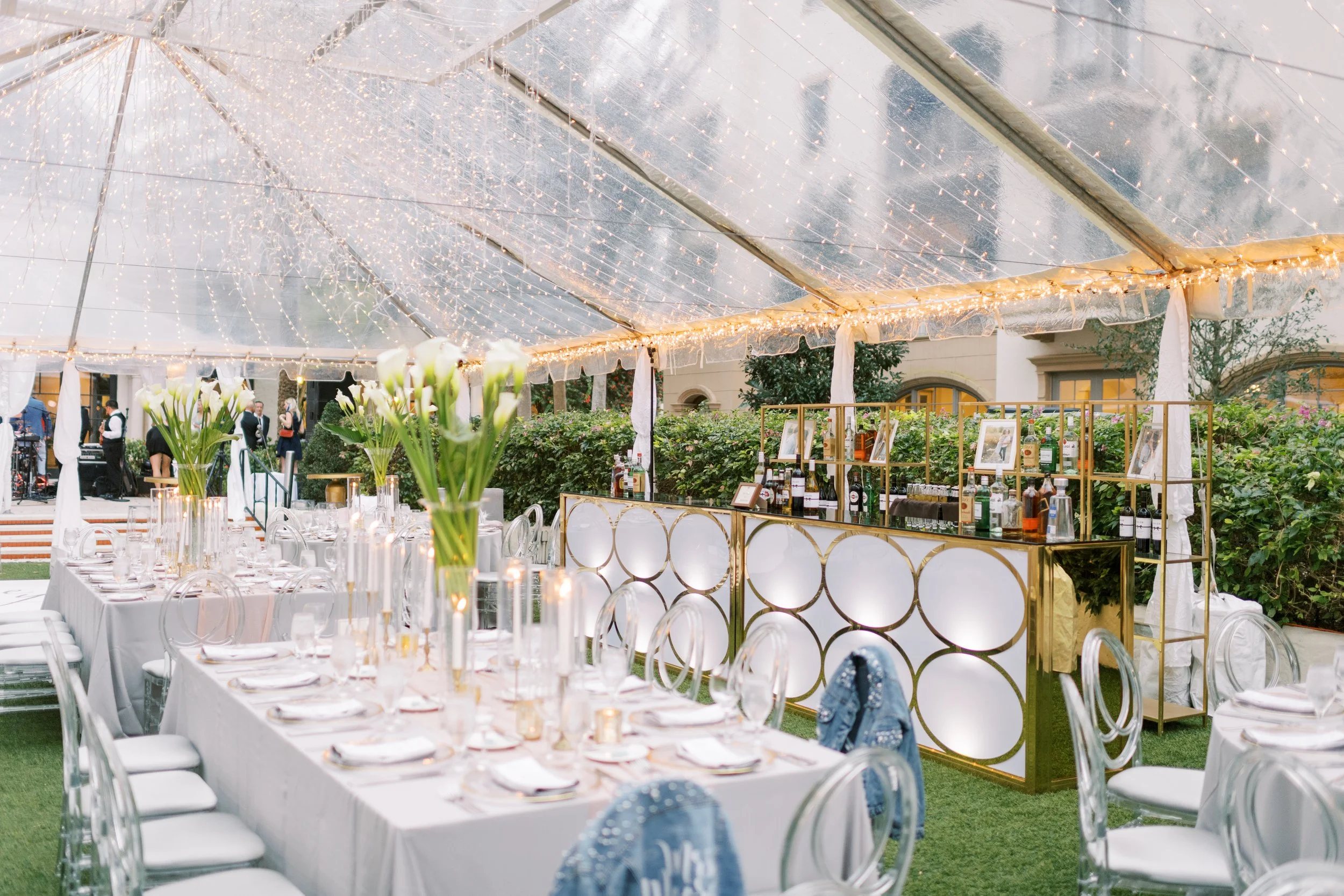 Elegant outdoor event space with tables set for a formal dinner under a clear tent decorated with string lights. The tables have white tablecloths, glassware, and floral centerpieces with white flowers. In the background, there is a bar with bottles of alcohol and glasses, and a lush garden.