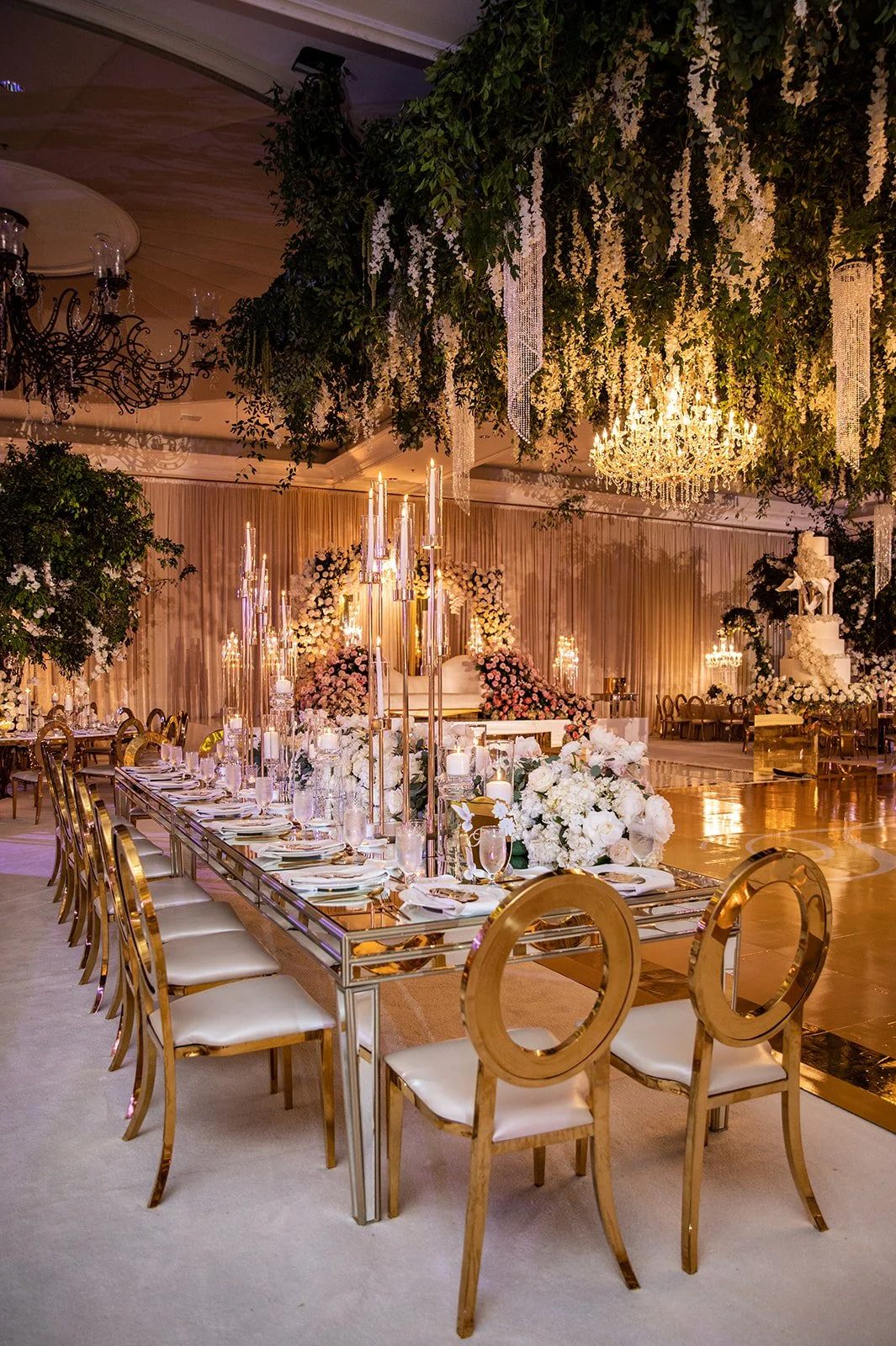 Elegant event space with a long table set for a celebration, decorated with white flowers, candles, and gold accents. Overhead is a lush floral and greenery arrangement with hanging crystals, and grand chandeliers illuminate the room.