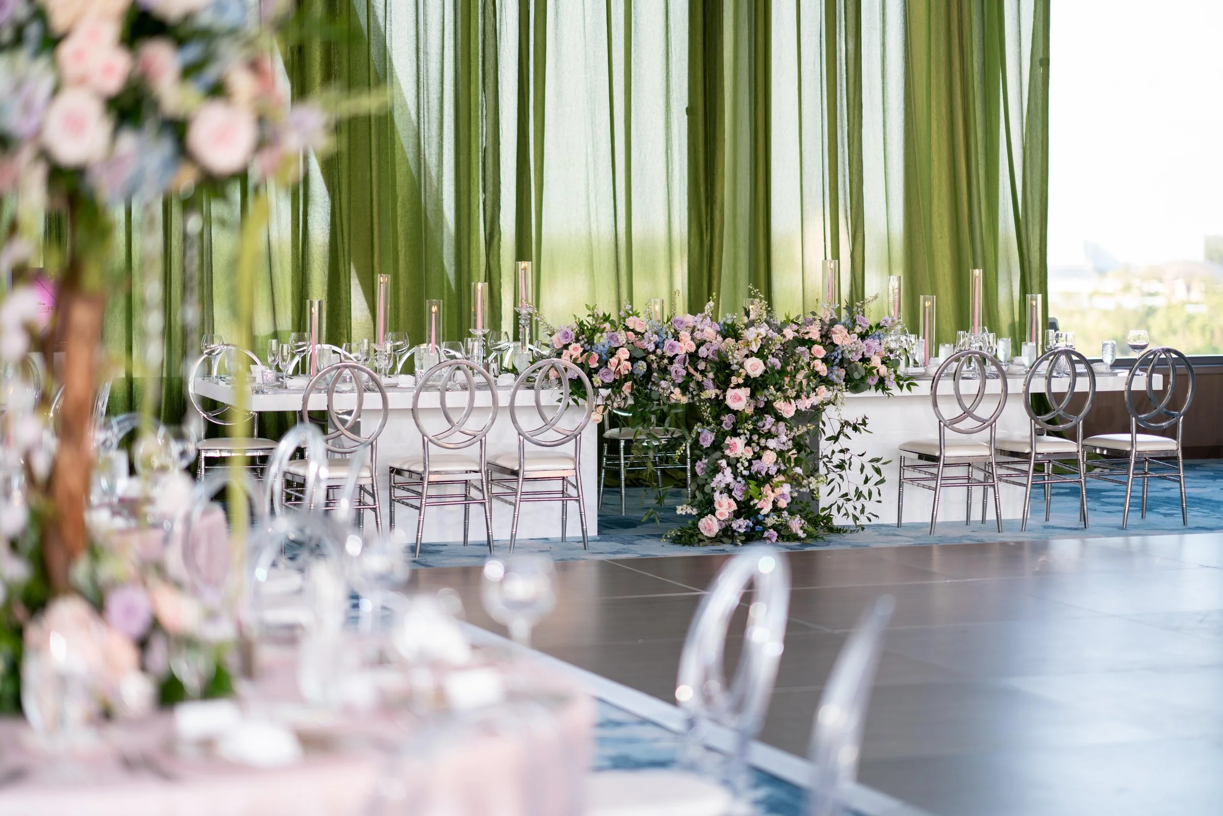 A beautifully decorated banquet table with pink and purple flowers and green foliage, set in front of large green curtains and a window with city views.