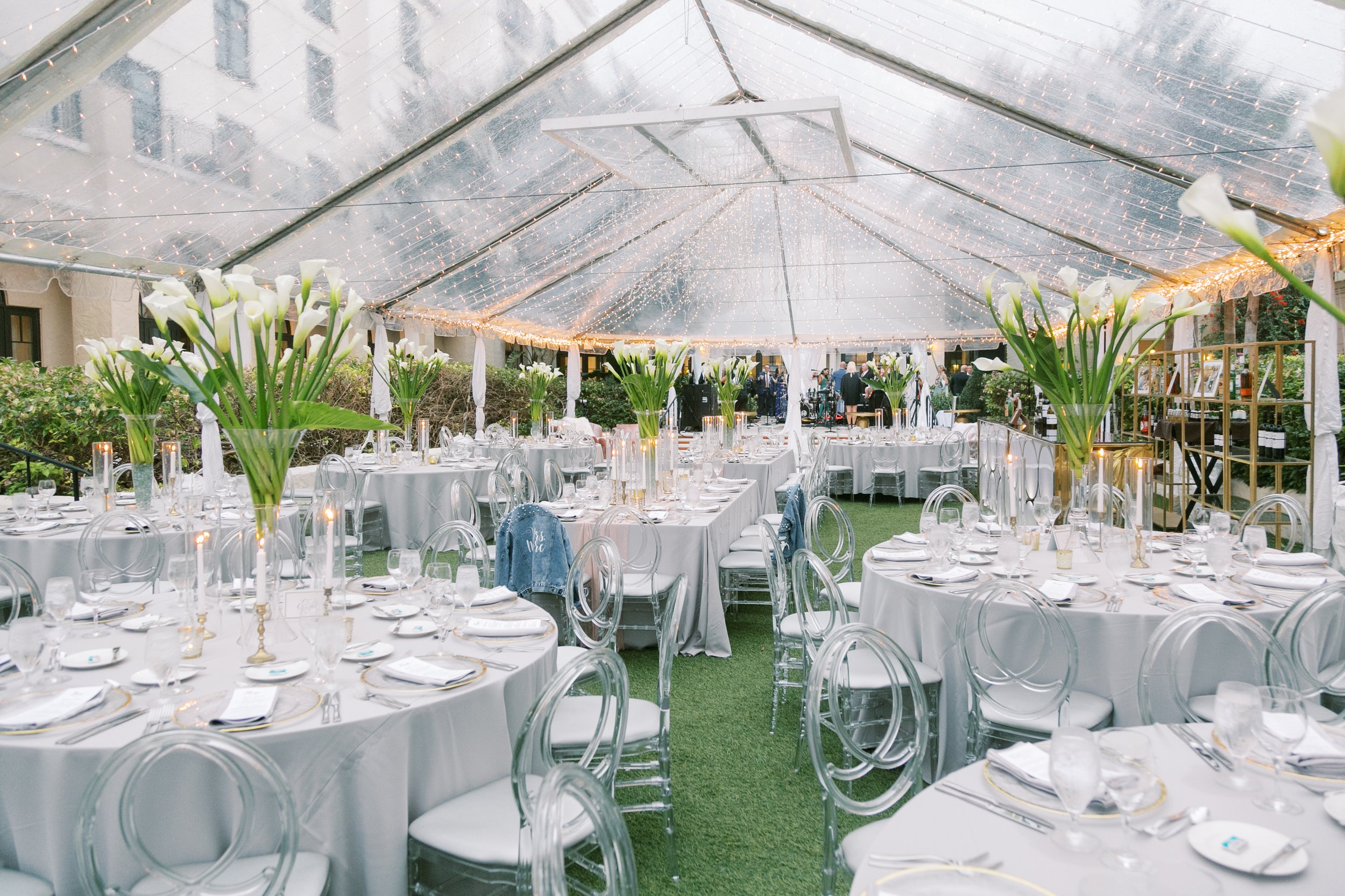 Elegant outdoor event setup with round tables covered in white tablecloths, decorated with tall vases of white calla lilies, set under a clear tent with string lights, and surrounded by transparent ghost chairs.