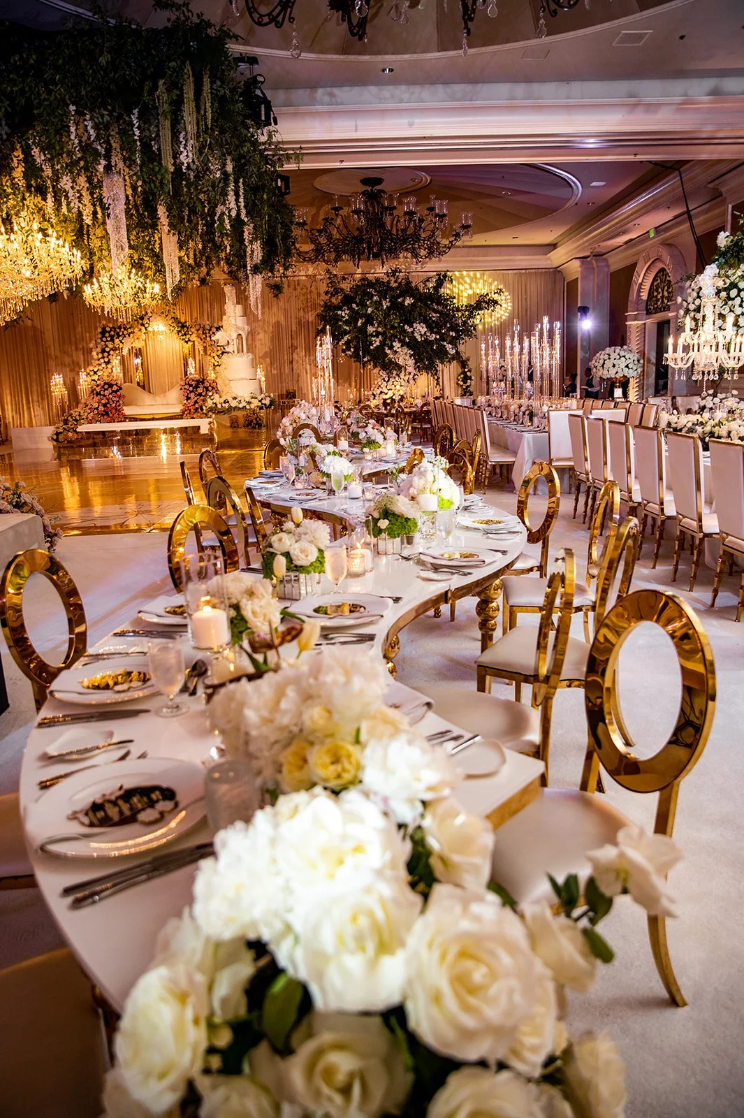 A lavishly decorated banquet hall with long tables set for a wedding reception, featuring white and gold accents, floral centerpieces, draped curtains, chandeliers, and a stage with a white sofa and floral arrangements.