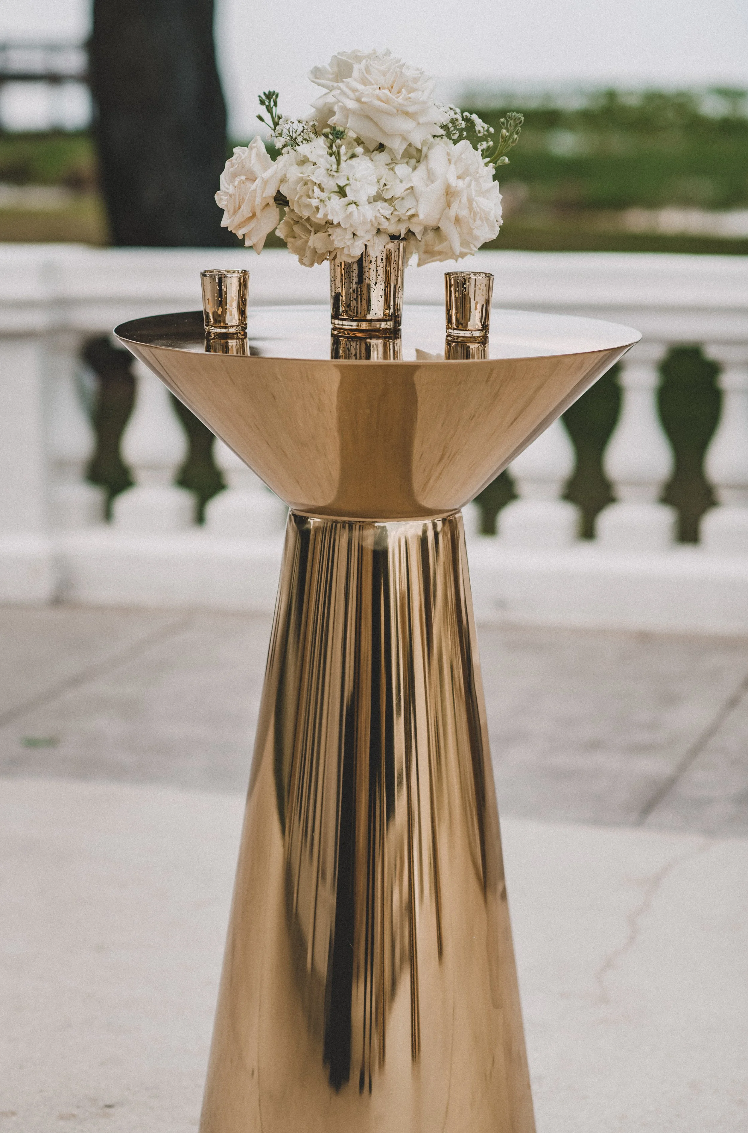 A tall, shiny, gold pedestal table with a bowl of white flowers, flanked by two small gold votive candle holders, on a terrace with a white balustrade in the background.