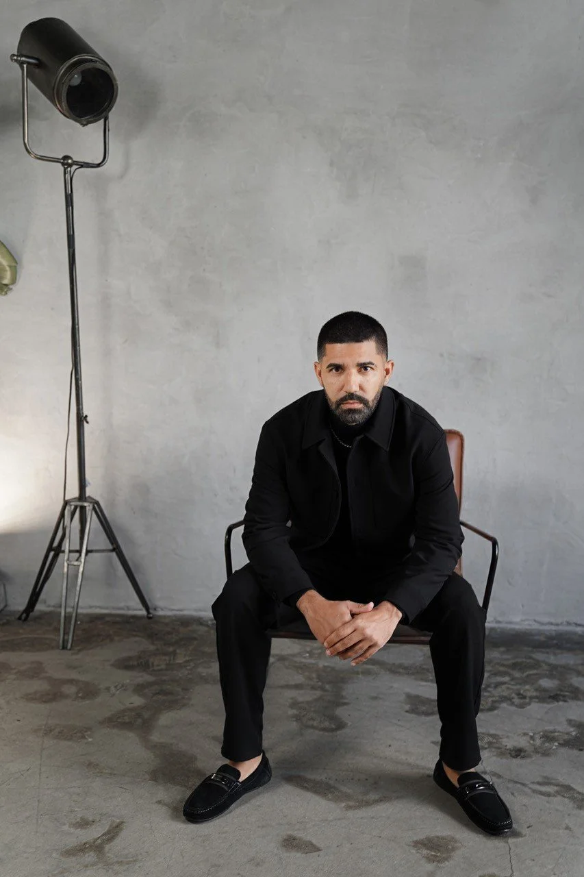 A man in all black seated on a chair in a studio with a plain gray wall and a vintage movie light on a stand to his left.