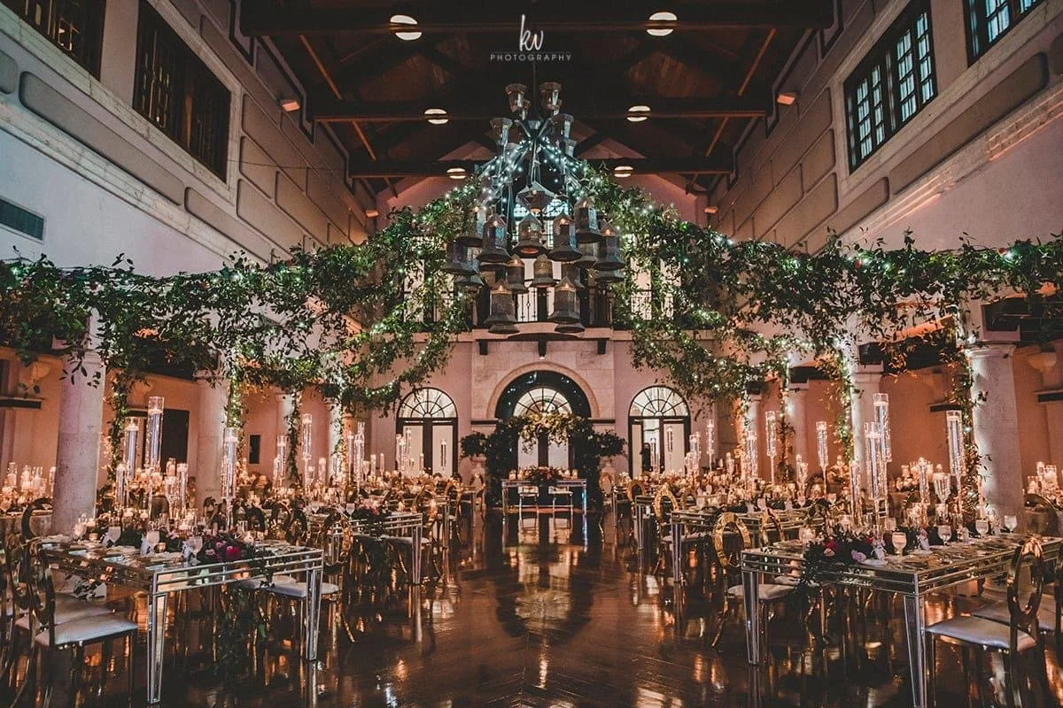 Elegant banquet hall decorated for a formal event with long tables, floral arrangements, candle centerpieces, and chandelier lighting.