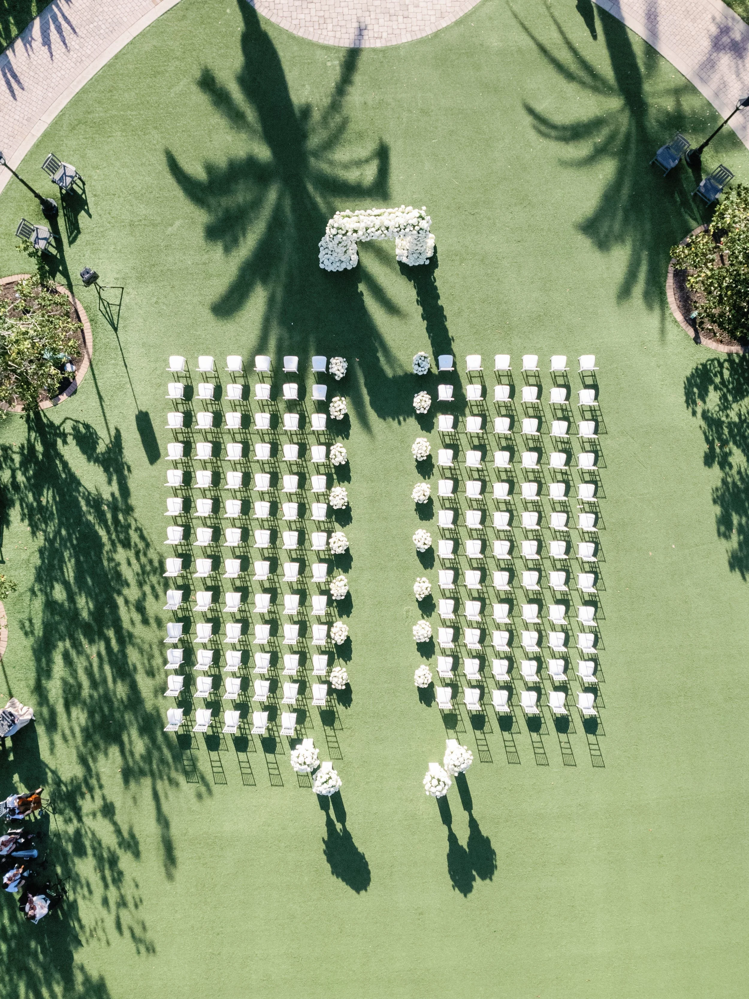 Empty outdoor wedding ceremony setup with rows of white chairs arranged on a green grass lawn, floral decorations, and large shadow of palm trees.