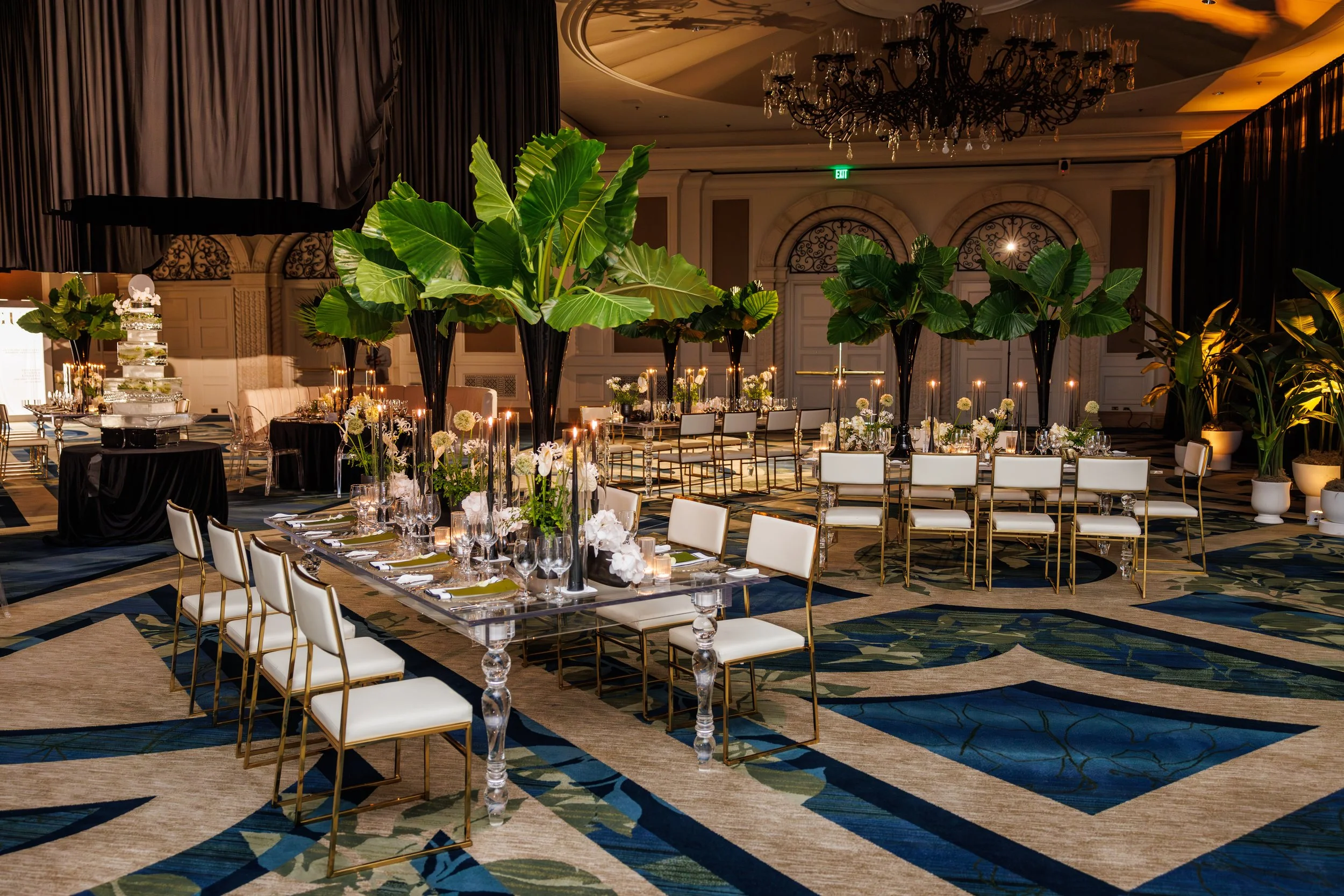 Elegant banquet setup in a luxurious ballroom with tall green tropical plants, white chairs with gold frames, and floral centerpieces, including white orchids and greenery, on a long dining table.