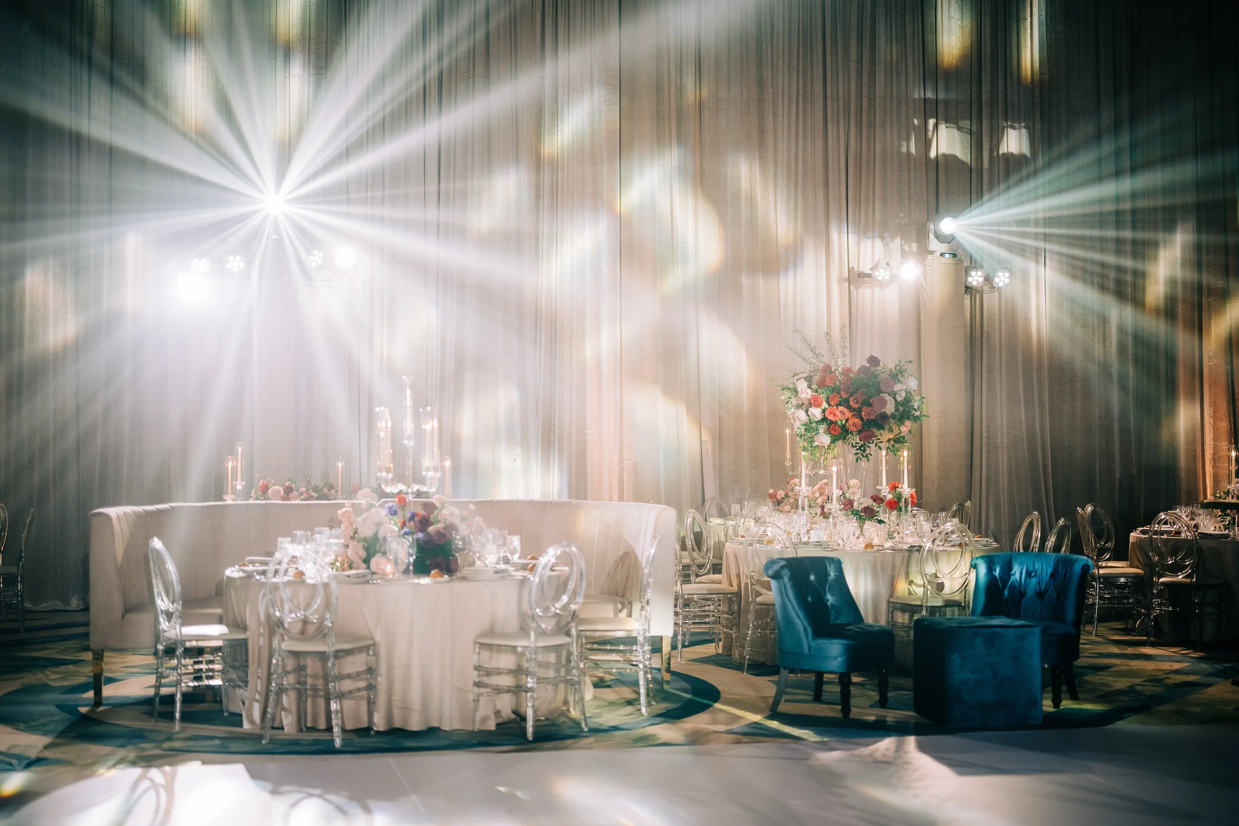Elegant banquet hall with round tables decorated with pink and red floral centerpieces, surrounded by transparent and blue chairs, with bright stage lights shining down on the scene.