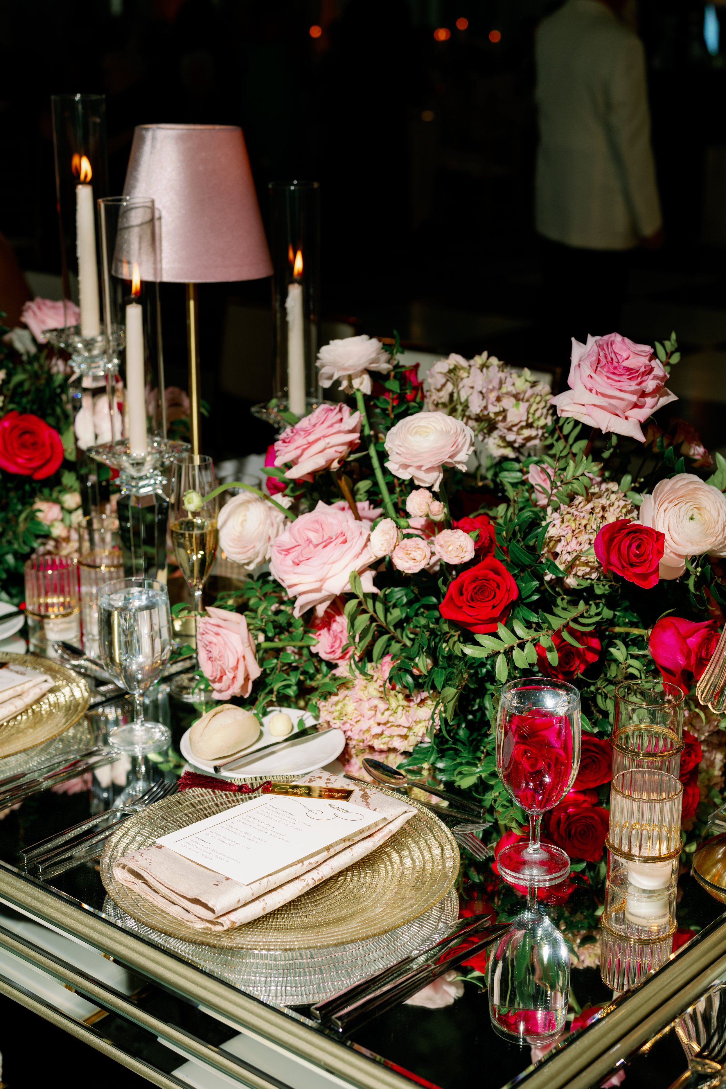 Elegant dining table decorated with pink and red roses, glassware, candles, and a menu, set for a formal event.