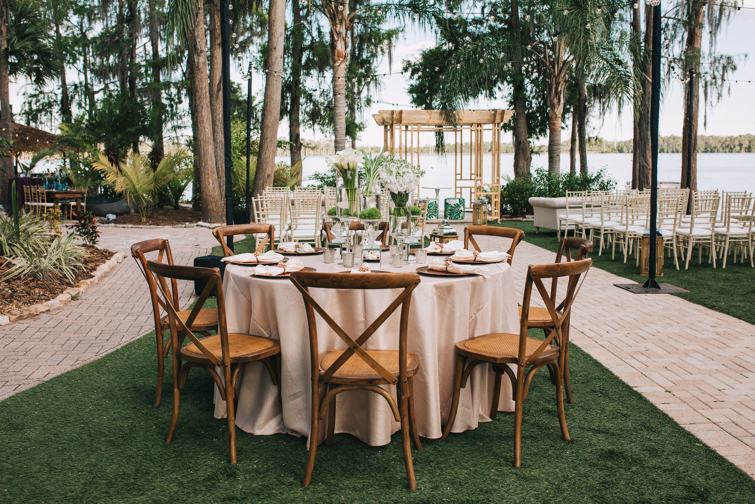 Round banquet table set for outdoor event with white tablecloth, floral centerpieces, and place settings, surrounded by wooden chairs, on a brick patio near water with trees and additional empty chairs in the background.