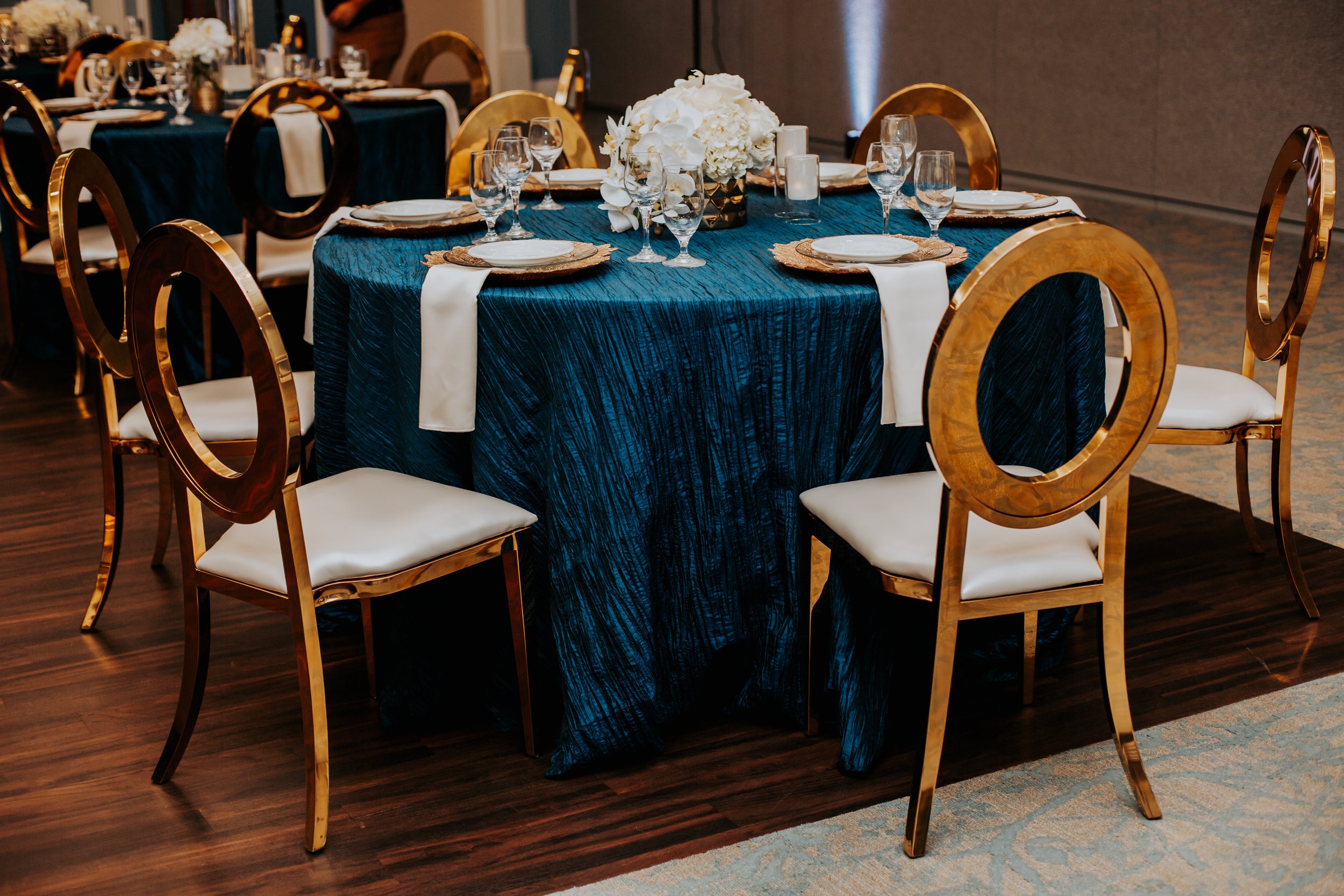 A round banquet table with a dark blue textured tablecloth, gold chairs with white cushions, and a centerpiece of white flowers in a gold vase, set with plates, napkins, glasses, and candles for an event or celebration.