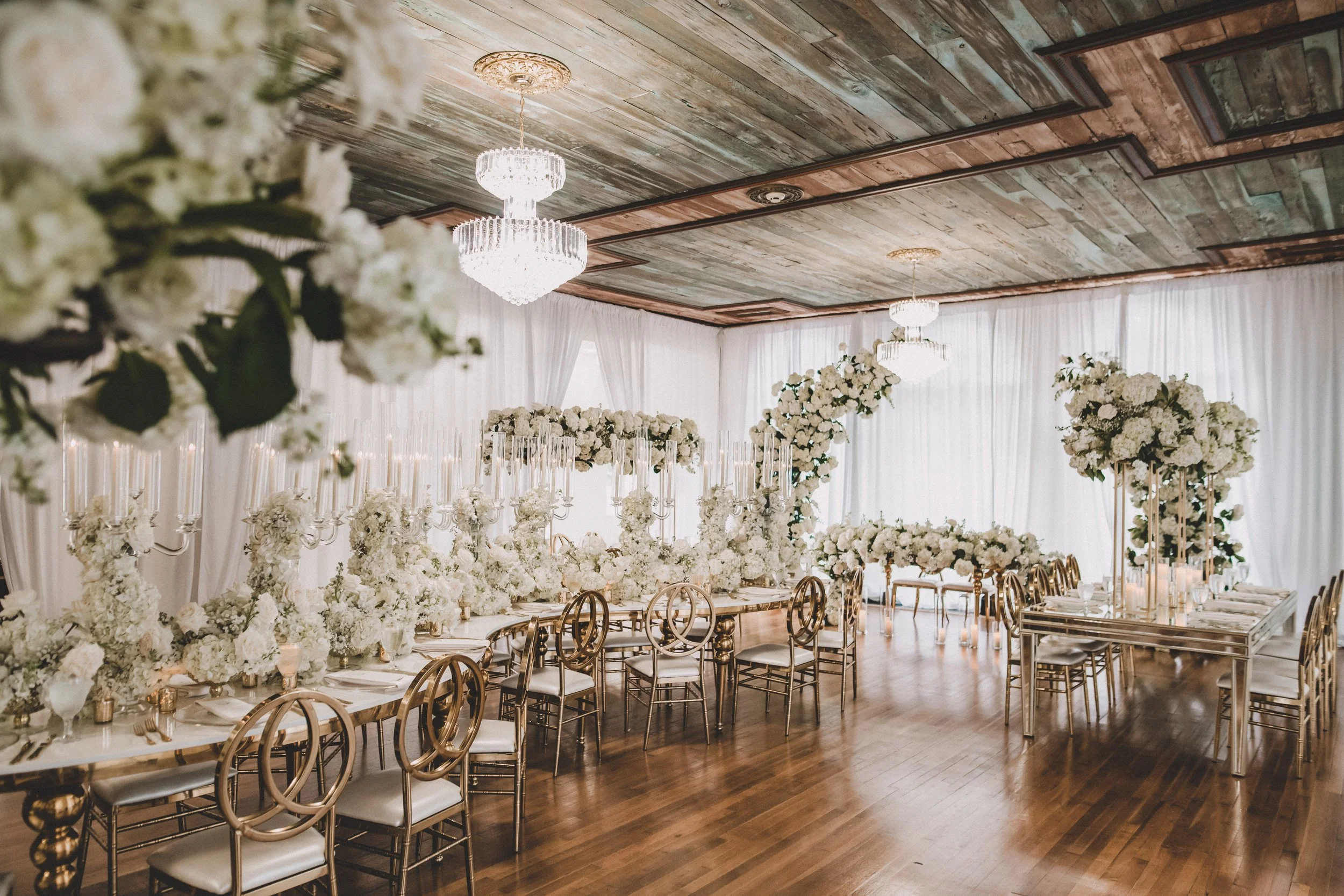 Elegant wedding reception decorated with white flowers, tall floral arrangements, chandeliers, and gold chairs in a spacious, well-lit room.