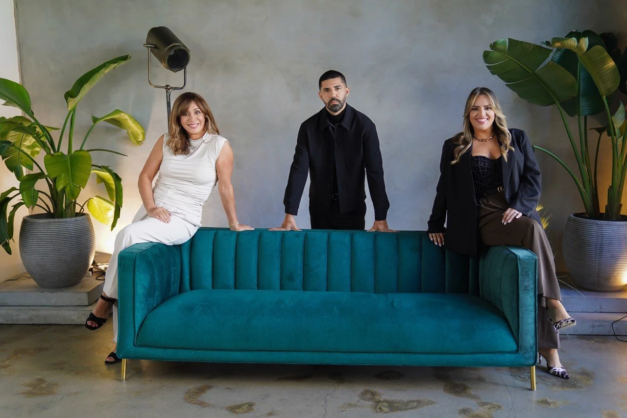 Three people, two women and one man, are posing behind and on a teal velvet sofa in a modern indoor space with large potted plants and a concrete floor.