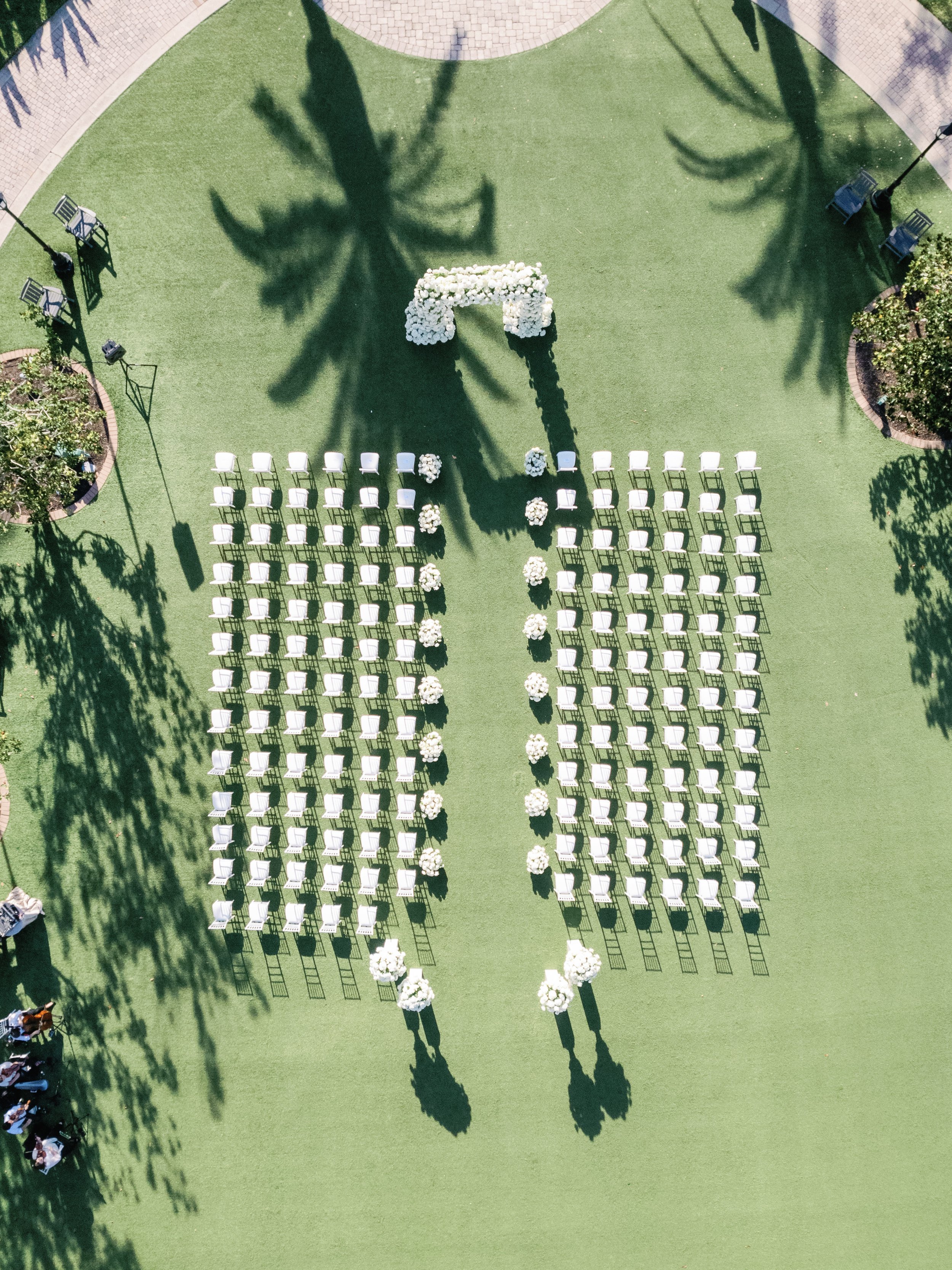 An outdoor wedding setup with white chairs arranged in rows on green grass, decorated with floral arrangements and an arch at the front, surrounded by trees casting shadows.